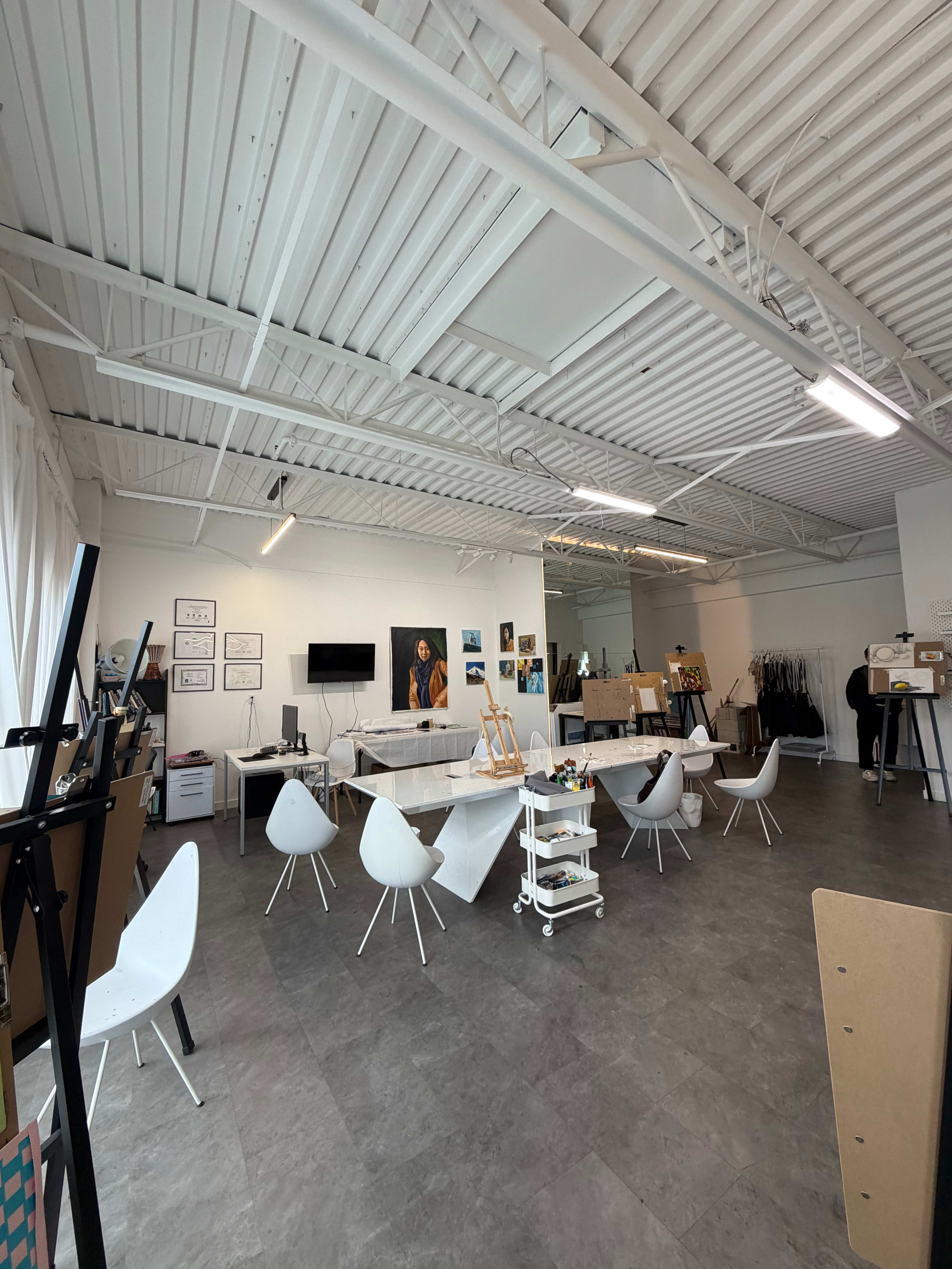 The image shows an art studio featuring several white chairs arranged around tables, with easels, artwork on the walls, and a large mirror.