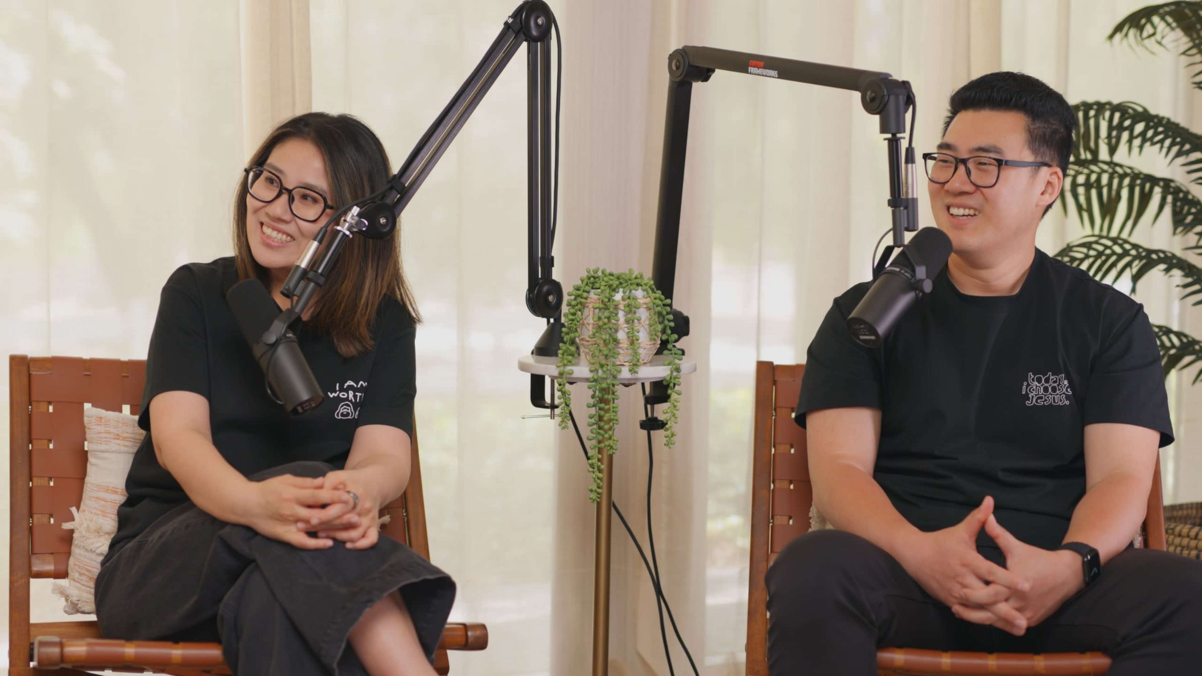 Two people sit in chairs, each beside a microphone on an adjustable stand, with a small green plant in the center of the scene.
