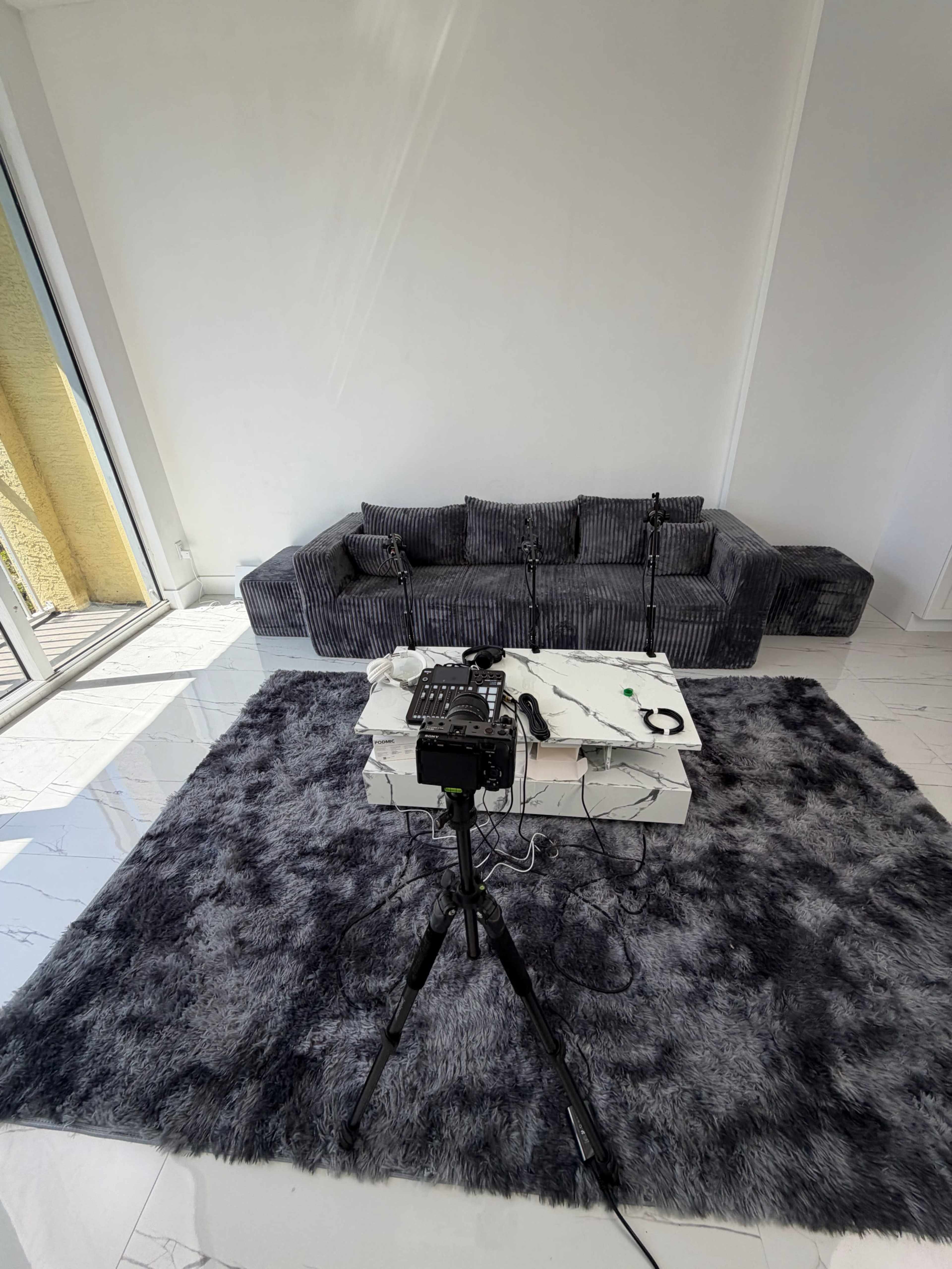 The image shows a modern living room with a gray sectional sofa, a coffee table on a gray fur rug, and a camera setup on a tripod facing the table.