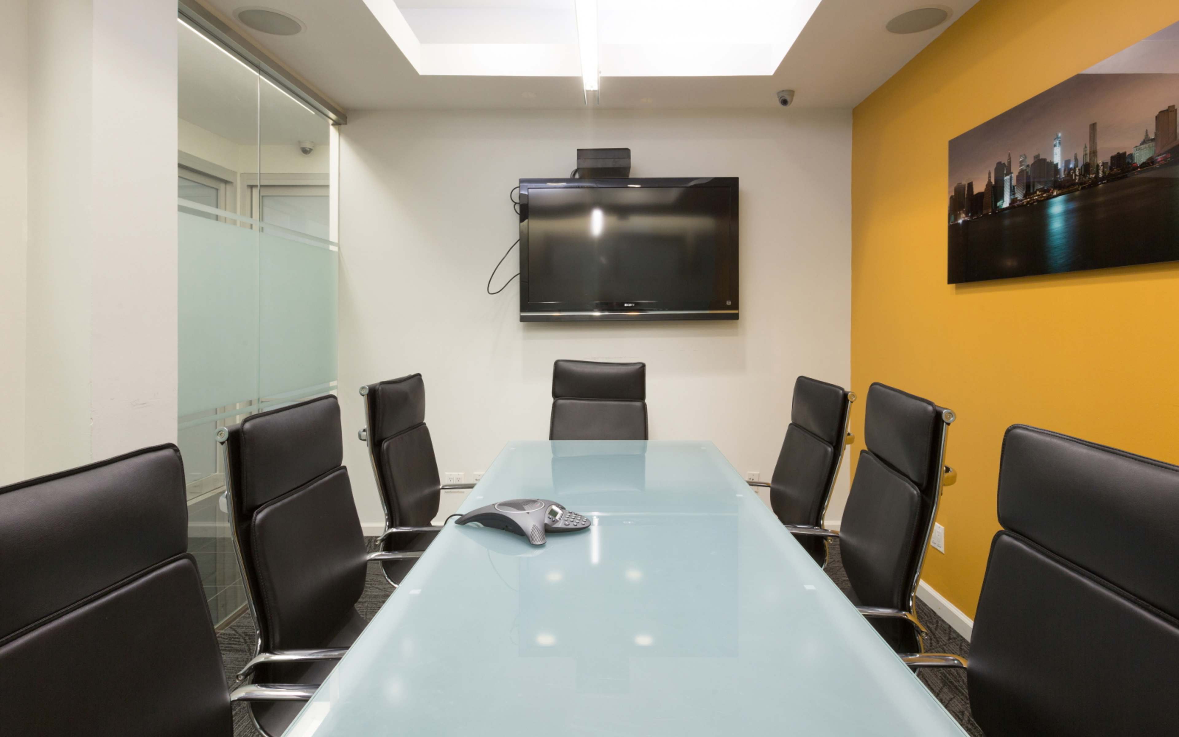 A modern conference room features a glass table surrounded by black chairs, with a television mounted on the wall and orange accent paint.