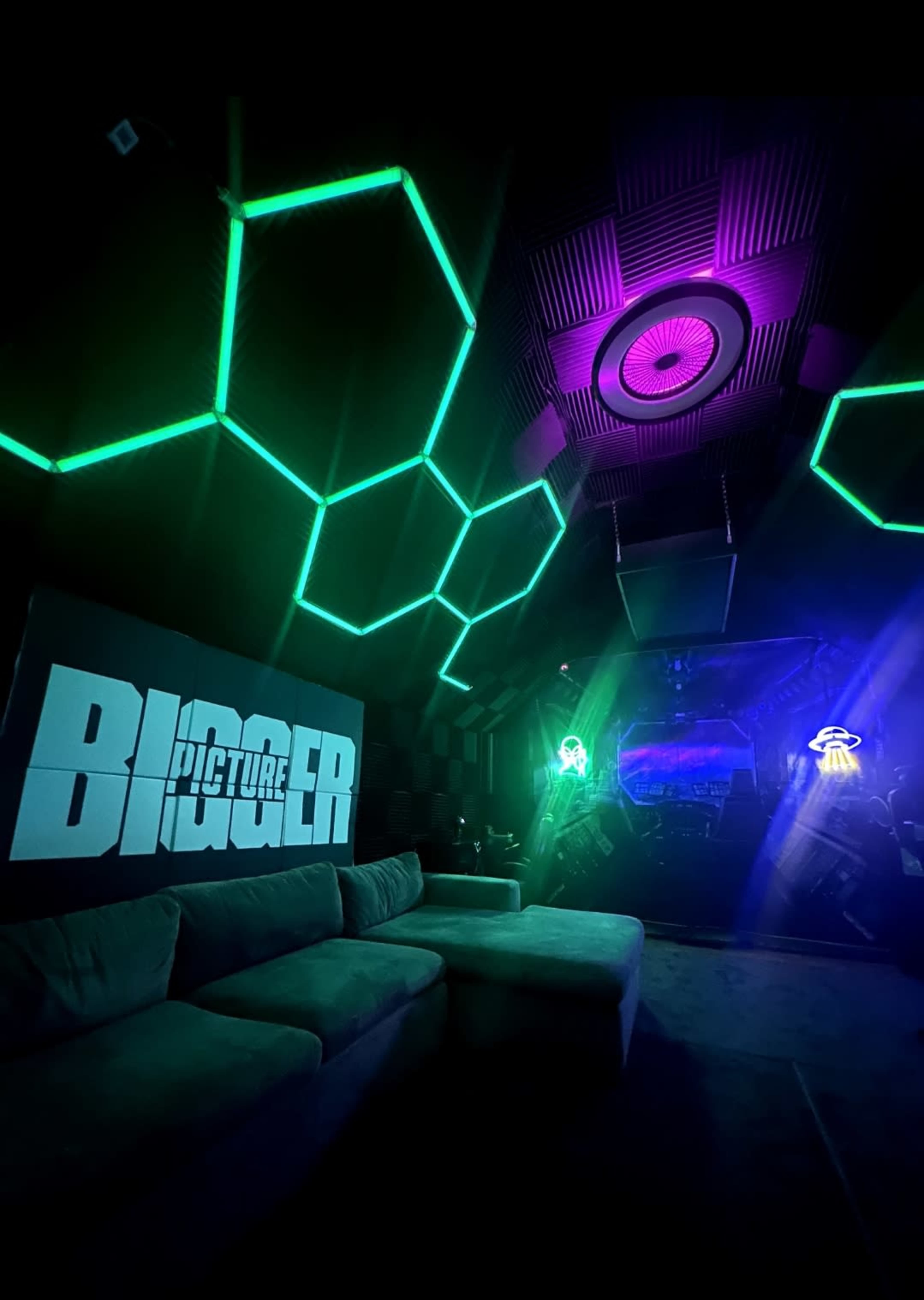 The image shows a modern room with hexagonal neon lights on the walls, a large couch, and a screen displaying the word "BIGGER."