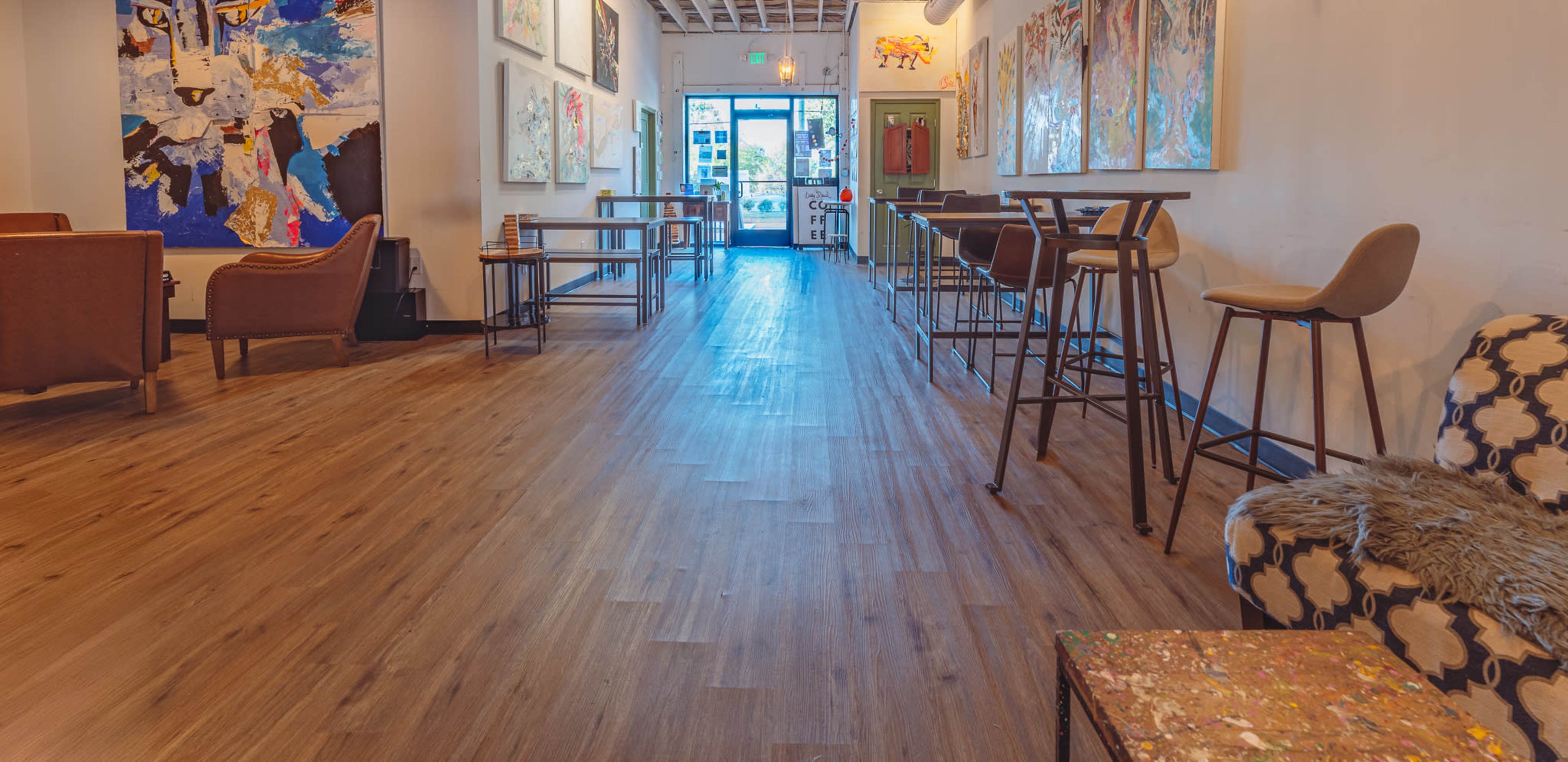 The image shows a narrow, well-lit hallway in a gallery or café, featuring artwork on the walls and furniture arranged on each side.