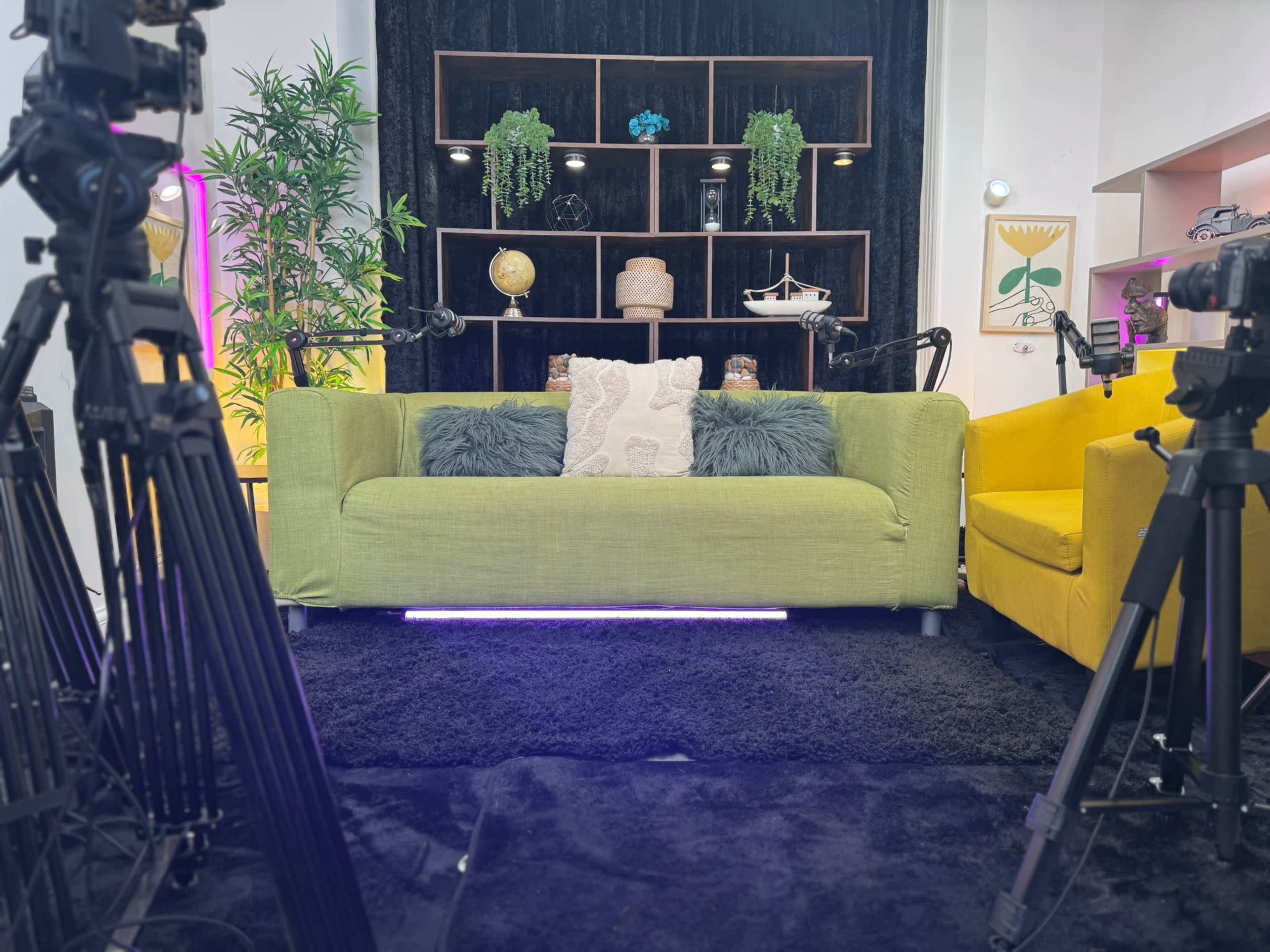 The image shows a brightly lit studio setup featuring a green sofa, a yellow armchair, and promotional decorations including plants and shelves in the background.