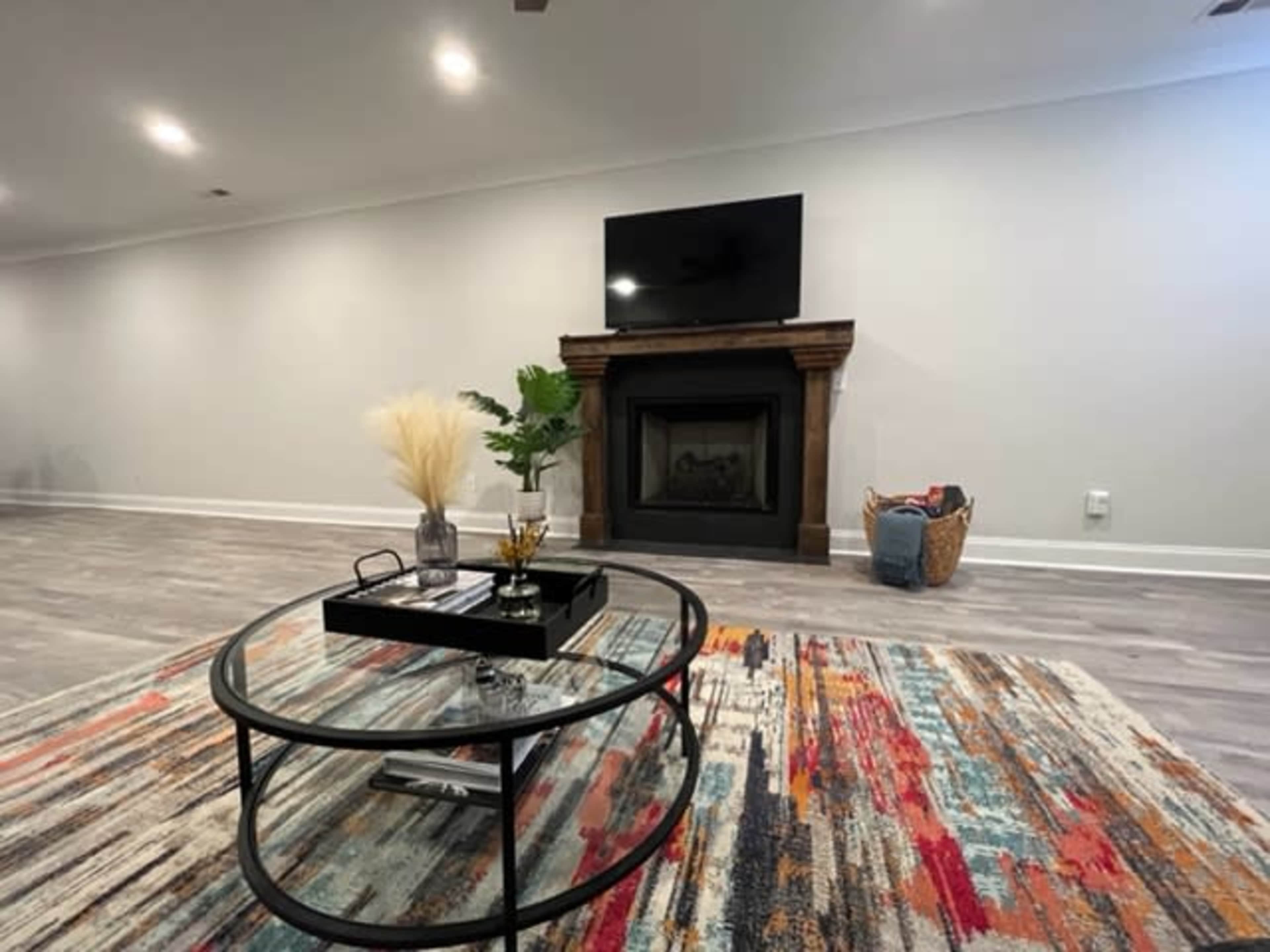 A modern living room featuring a round glass coffee table, a colorful area rug, a fireplace with a TV mounted above it, and a plant on the table.