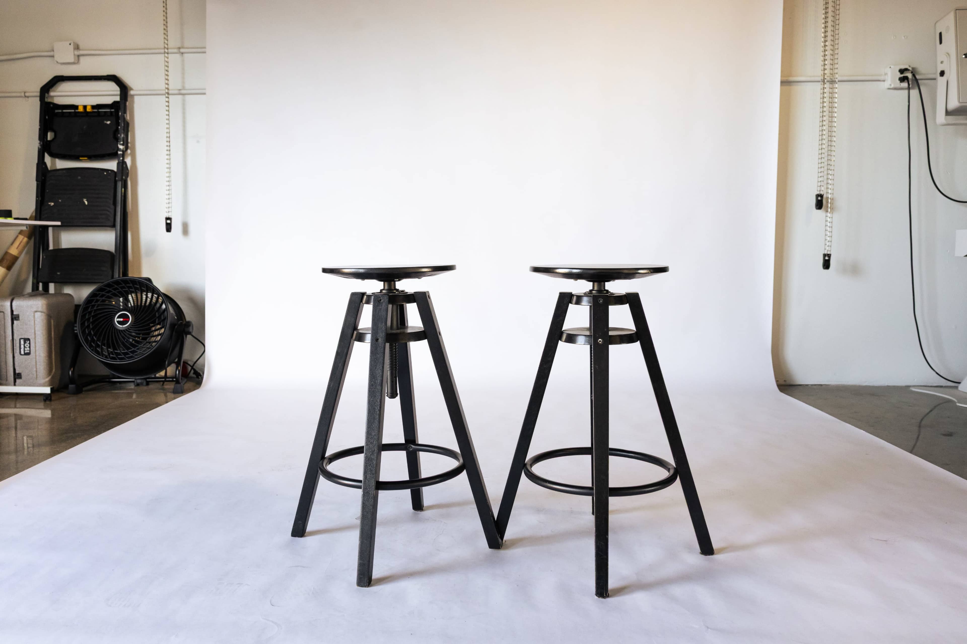 Two black metal bar stools with circular seats are positioned on a white backdrop in a well-lit interior space.