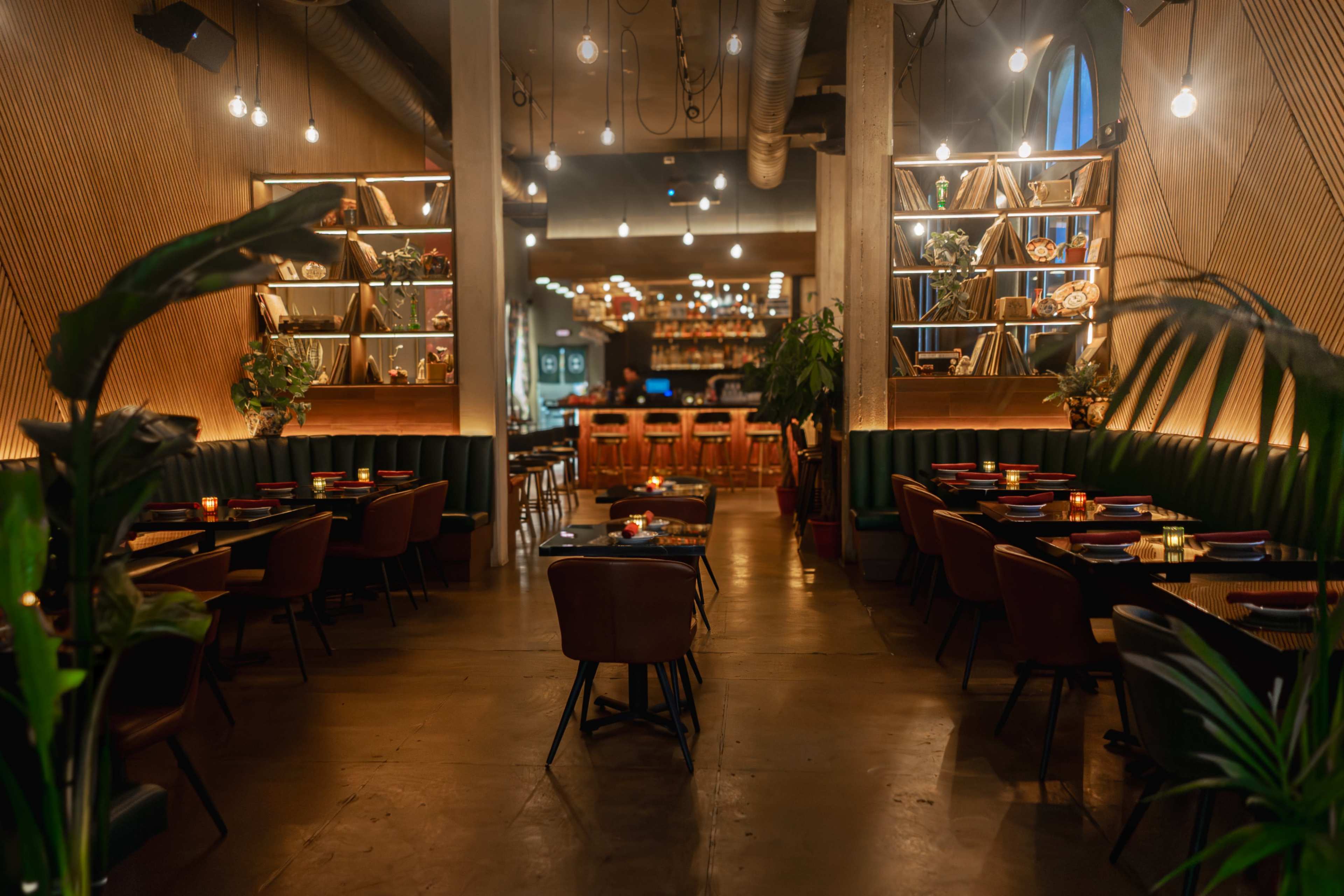 The image shows a modern restaurant interior featuring wooden shelves with decor, intimate seating arrangements with tables, and warm lighting fixtures.