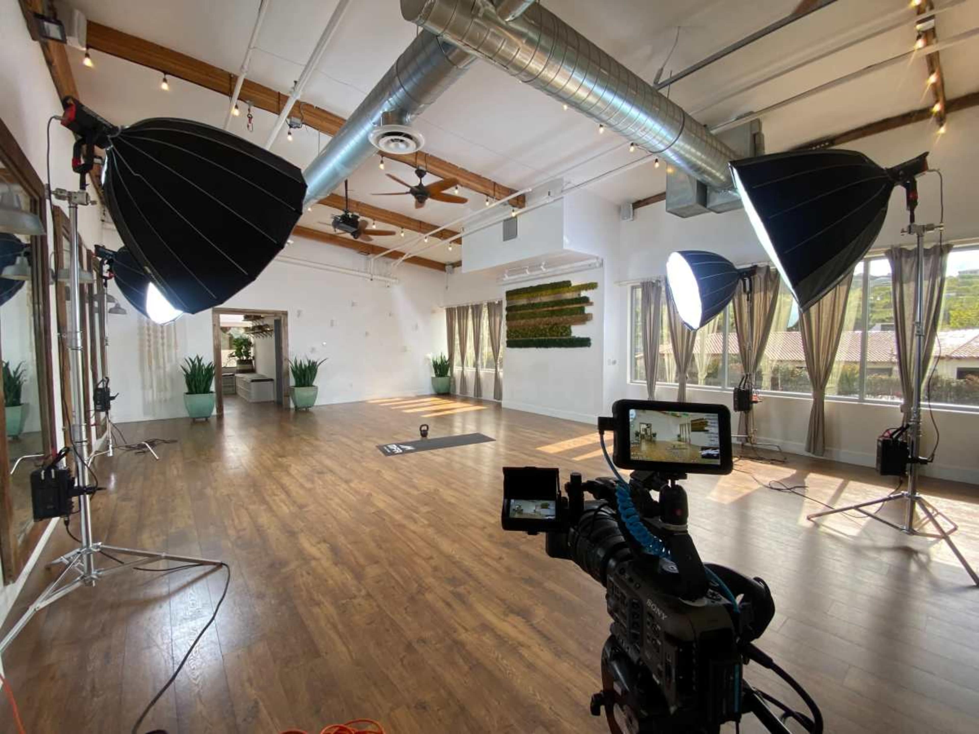 The image shows a spacious photography studio with large softbox lights, a camera on a tripod, and a wooden floor.