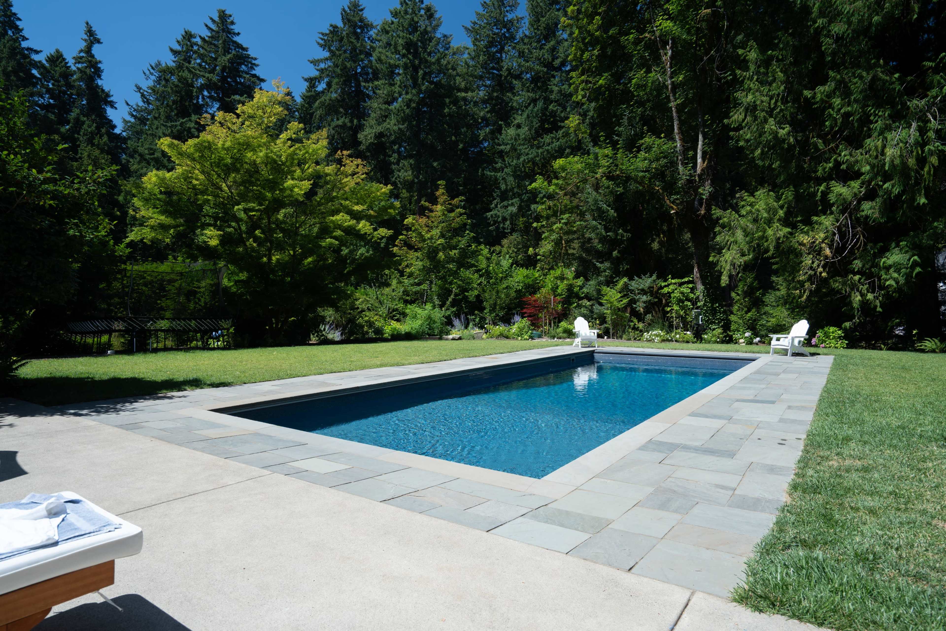 A rectangular swimming pool is surrounded by a stone patio and lush green landscaping, with tall trees in the background.