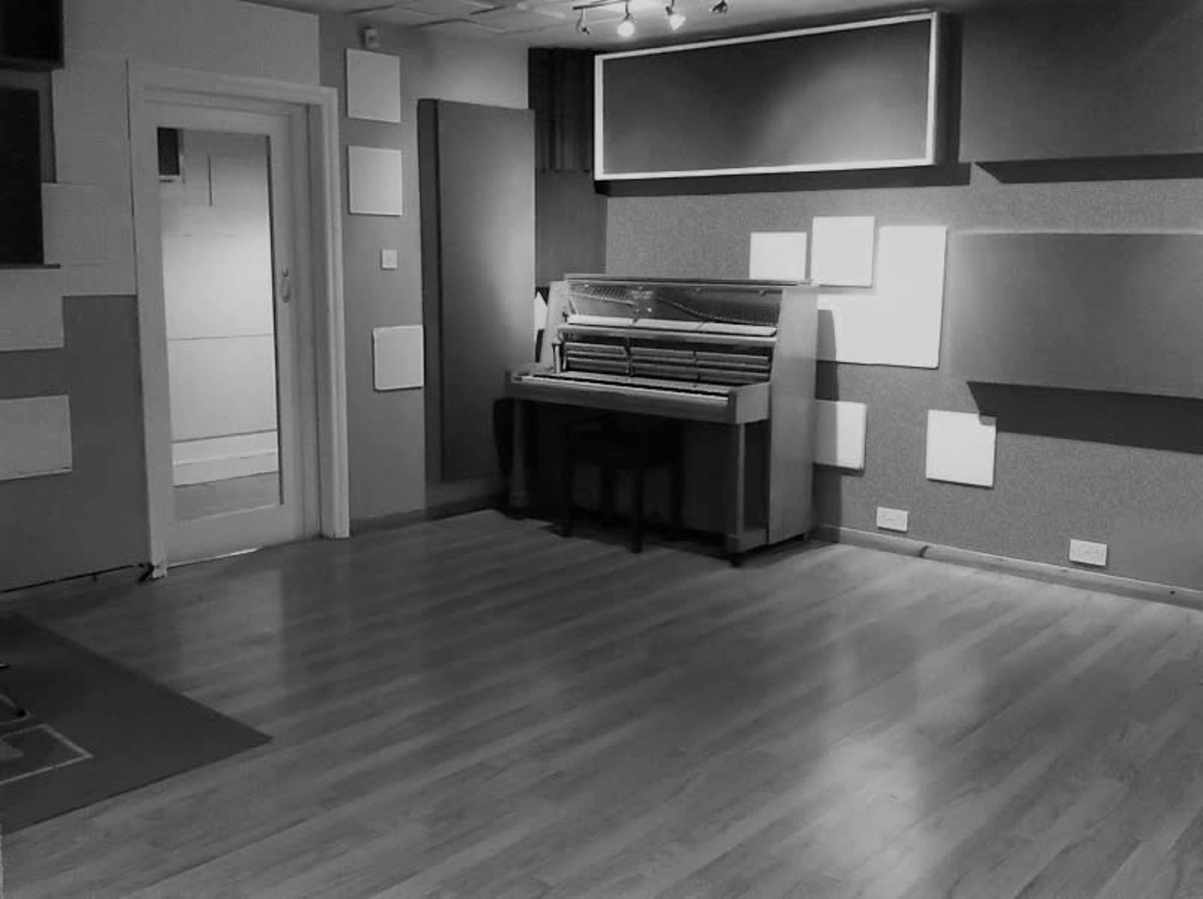 A music studio with a piano placed against a wall, surrounded by soundproofing panels and a doorway leading to another room.