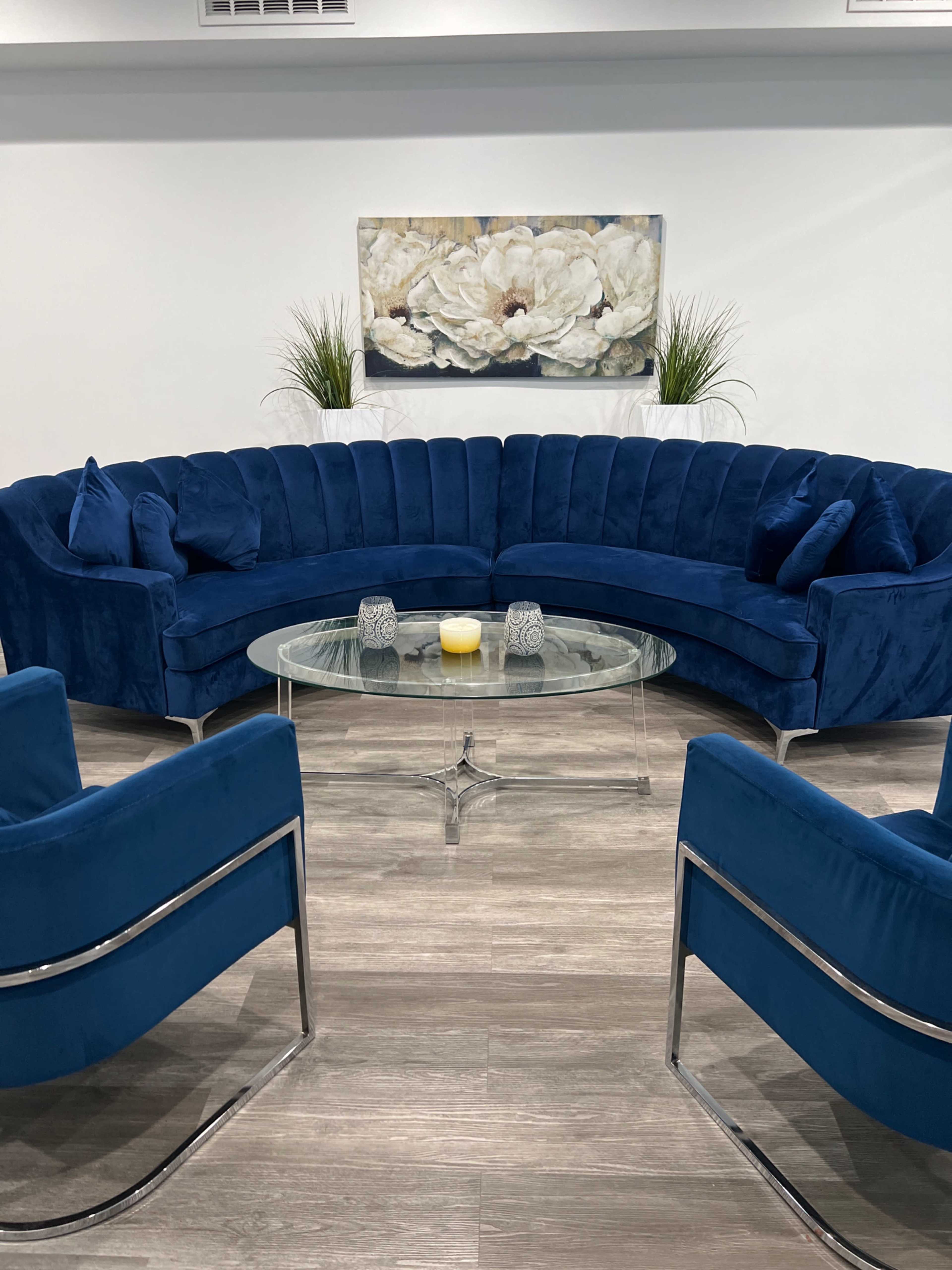 A modern lounge area with a curved blue velvet sofa, two matching armchairs, a glass coffee table, and a decorative painting of white flowers on the wall.