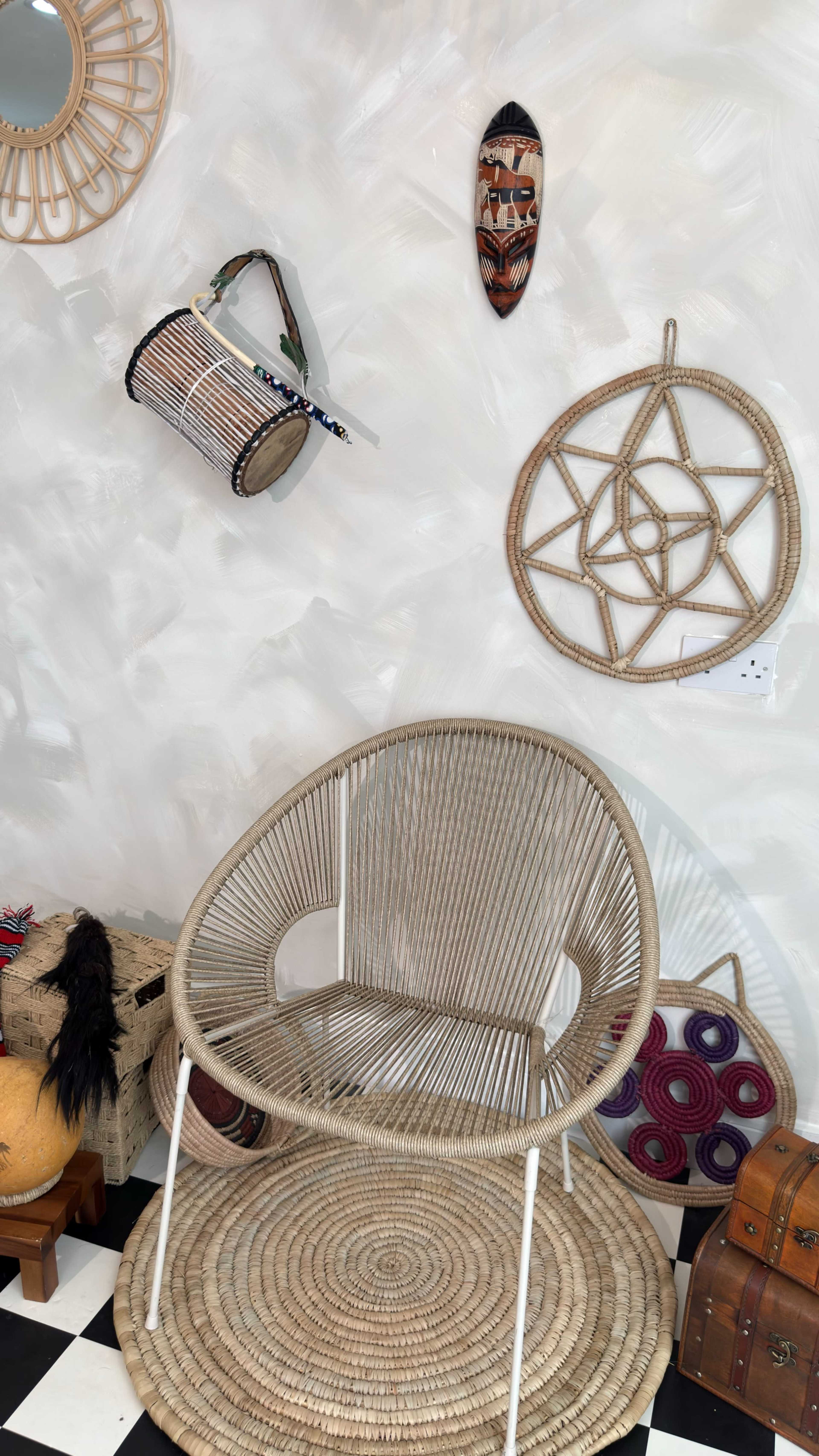 The image features a stylish rattan chair positioned on a round woven rug, surrounded by various decorative items including wall masks, a woven basket, and colorful textiles.