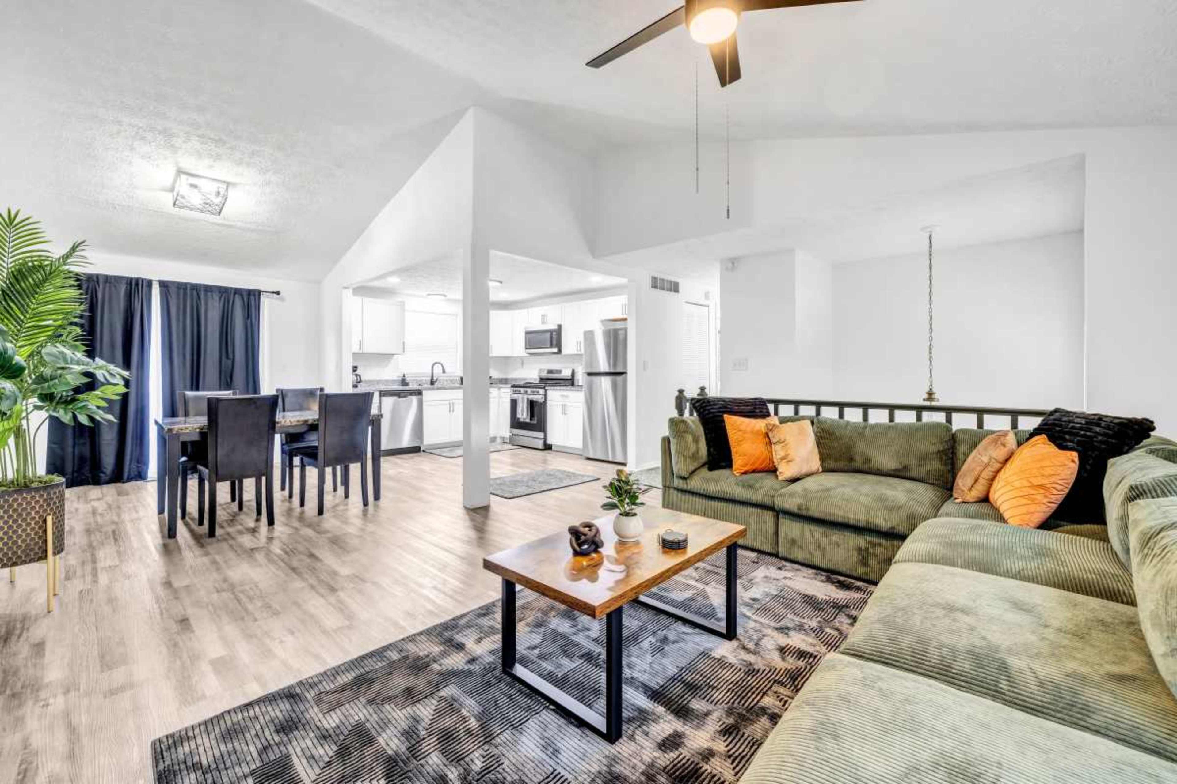 The image shows a modern living space featuring a green sectional sofa, a wooden coffee table, a dining area with a black table, and a bright kitchen in the background.