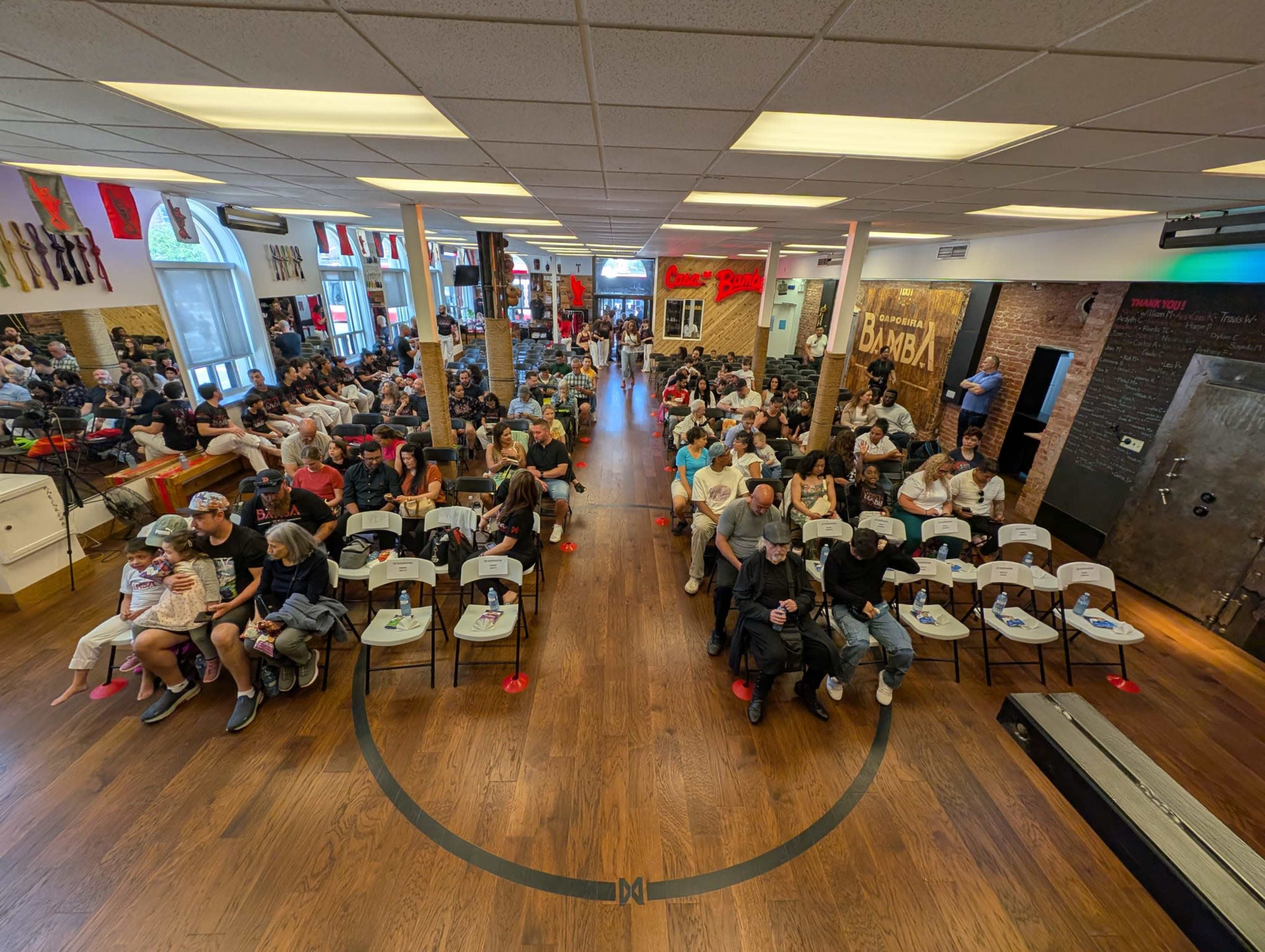 A large crowd of people is seated in a spacious room with wooden flooring, waiting for an event at a venue decorated with various flags and artistic wall designs.