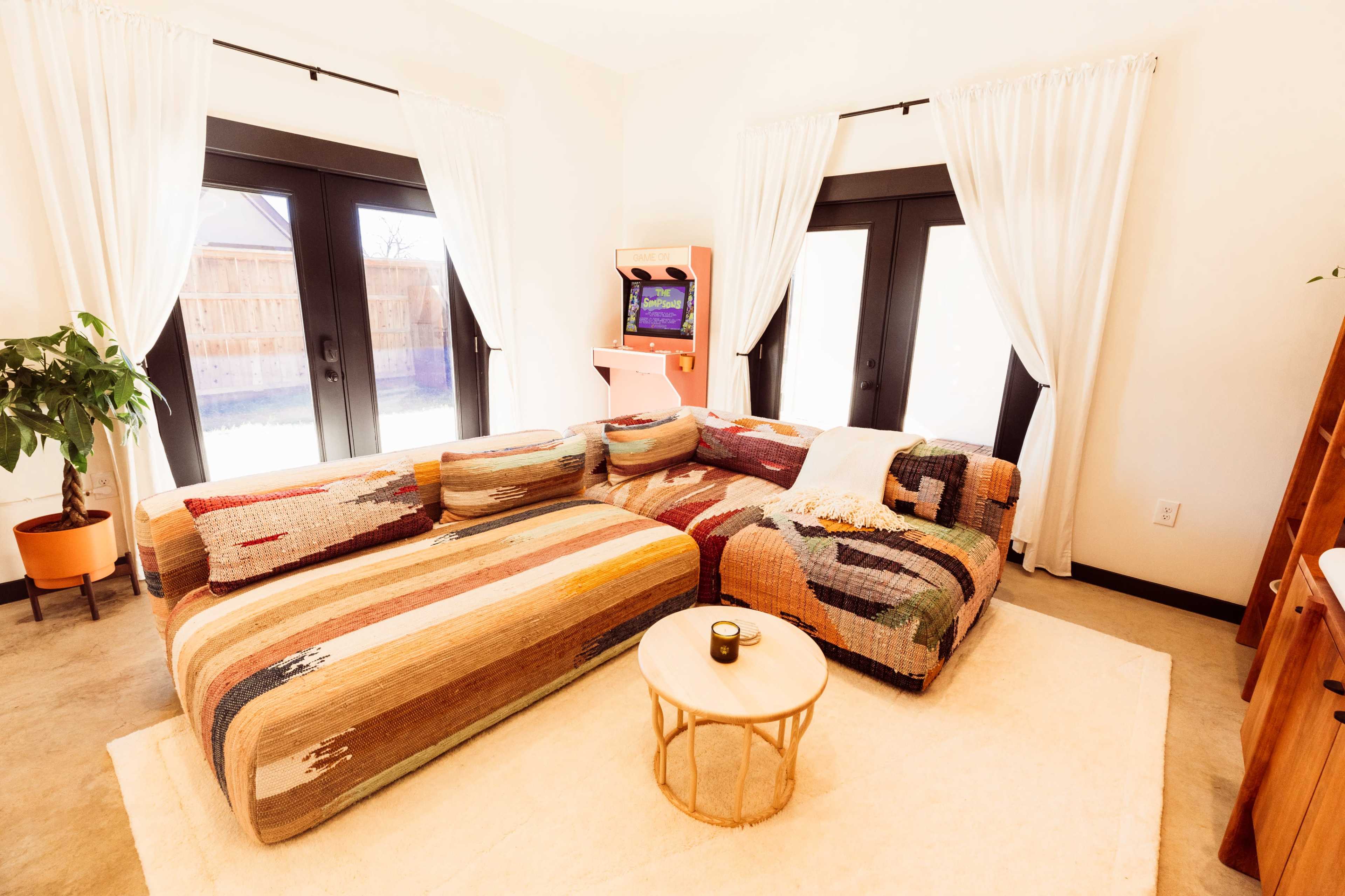 A brightly lit living room with a colorful sectional sofa, a round coffee table, and large windows facing an outdoor area.