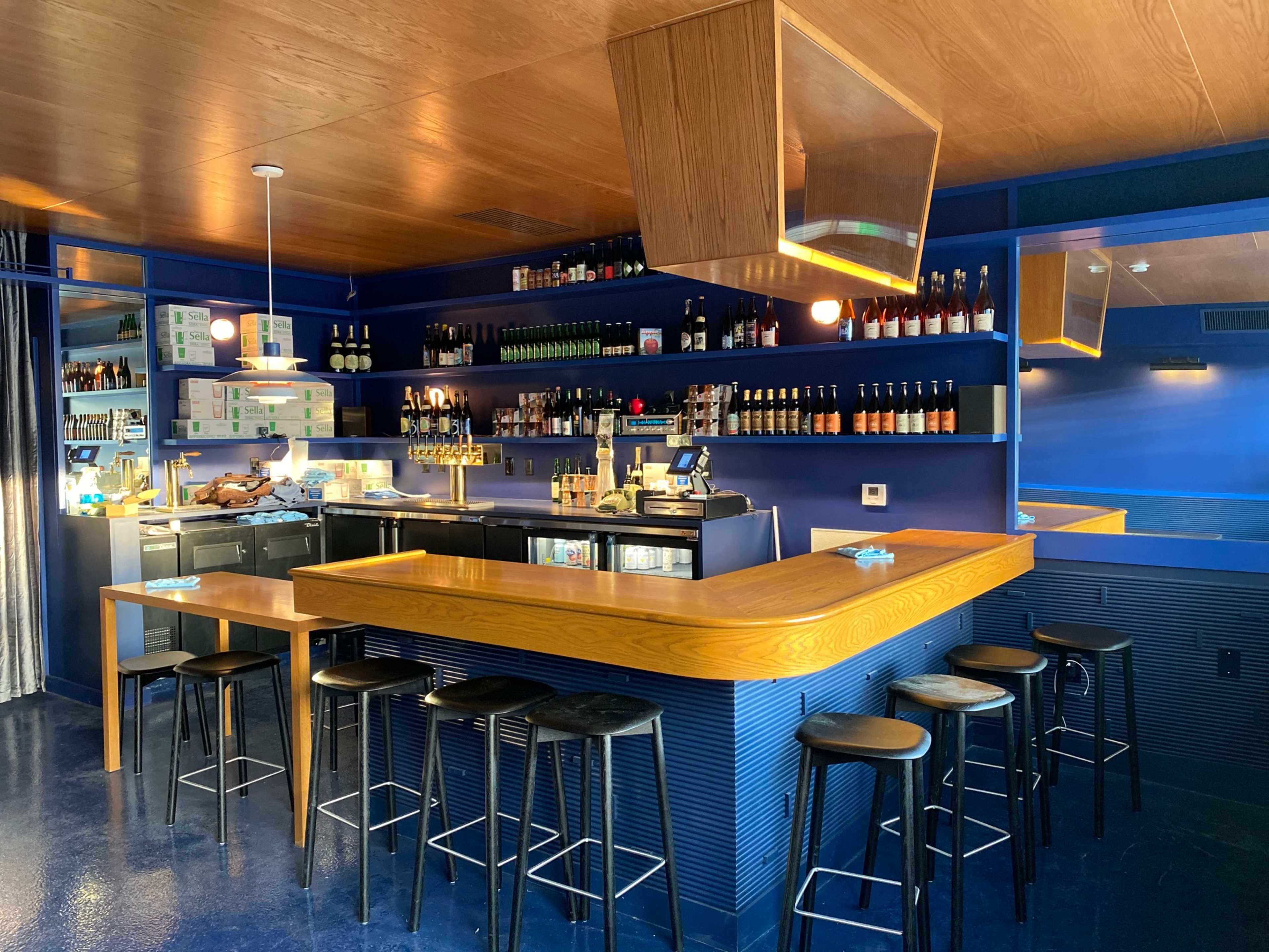 A modern bar area with a wooden countertop, black stools, and shelves lined with bottles against a blue wall.