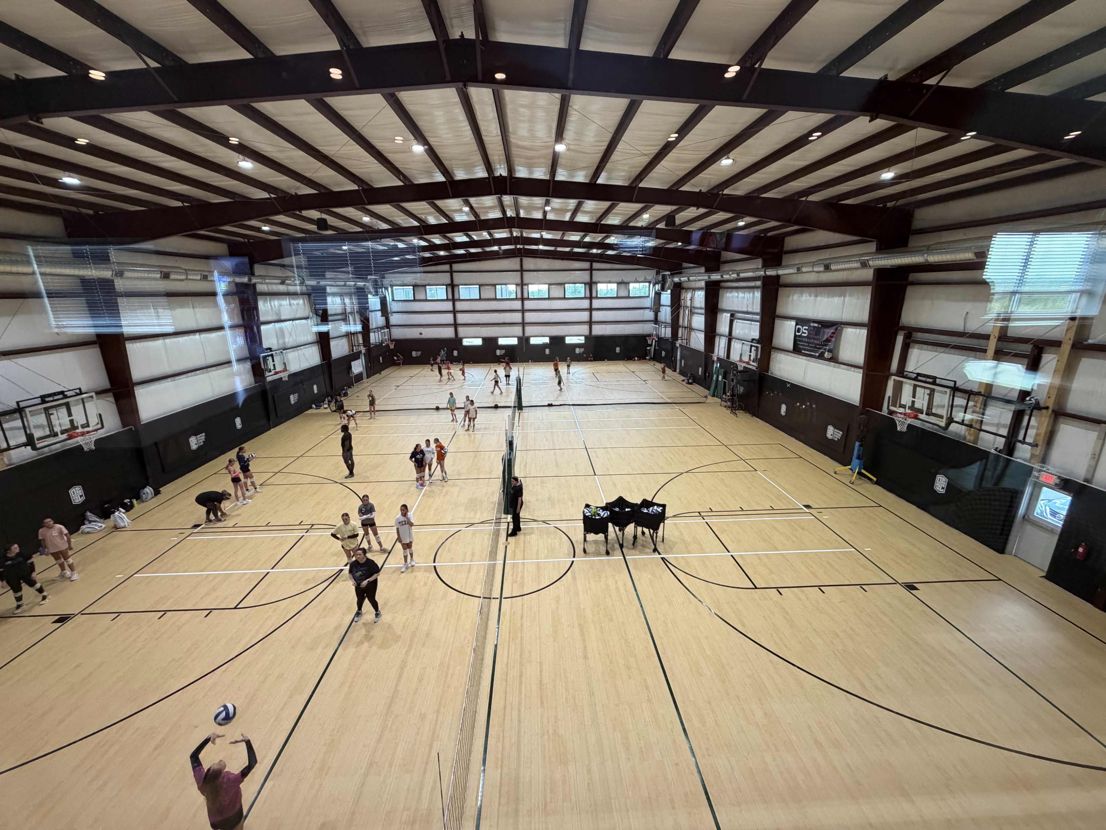 The image shows a large indoor sports facility with multiple volleyball courts and groups of people engaged in various activities.