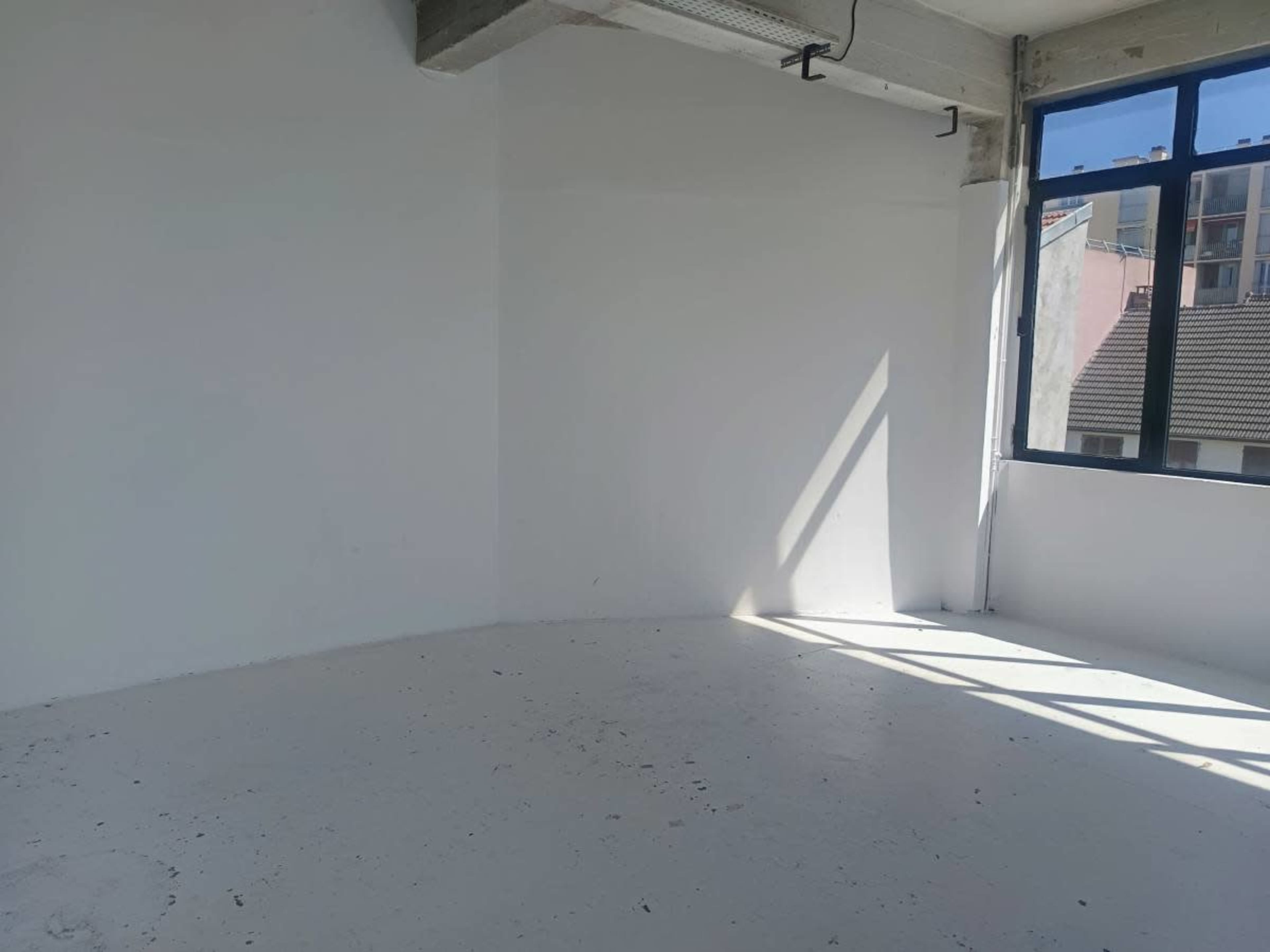 The image shows a bright, empty room with white walls and a large window allowing natural light to enter.
