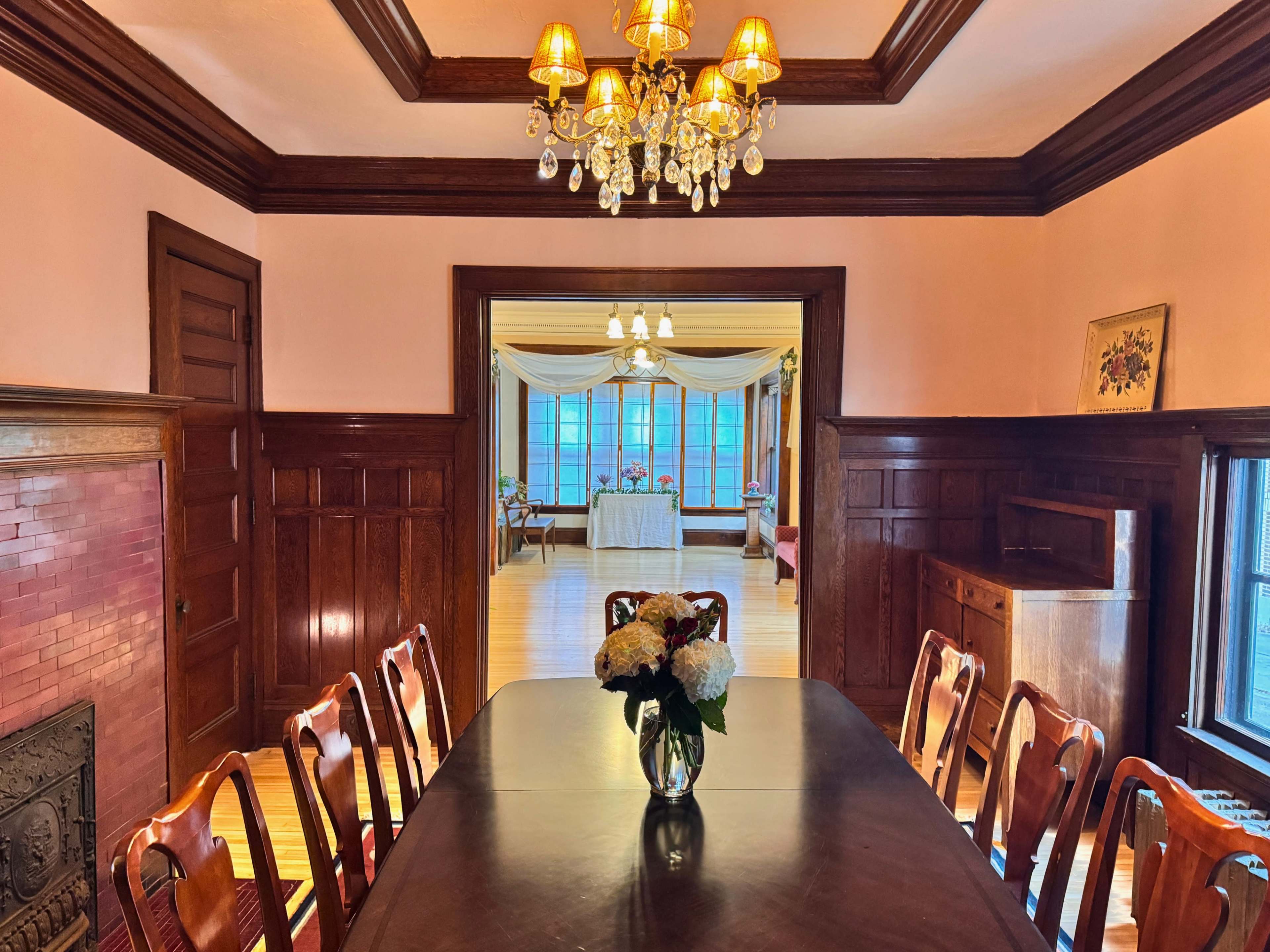 A large dining room features a long wooden table set with a vase of flowers, framed by paneled walls and a chandelier above.