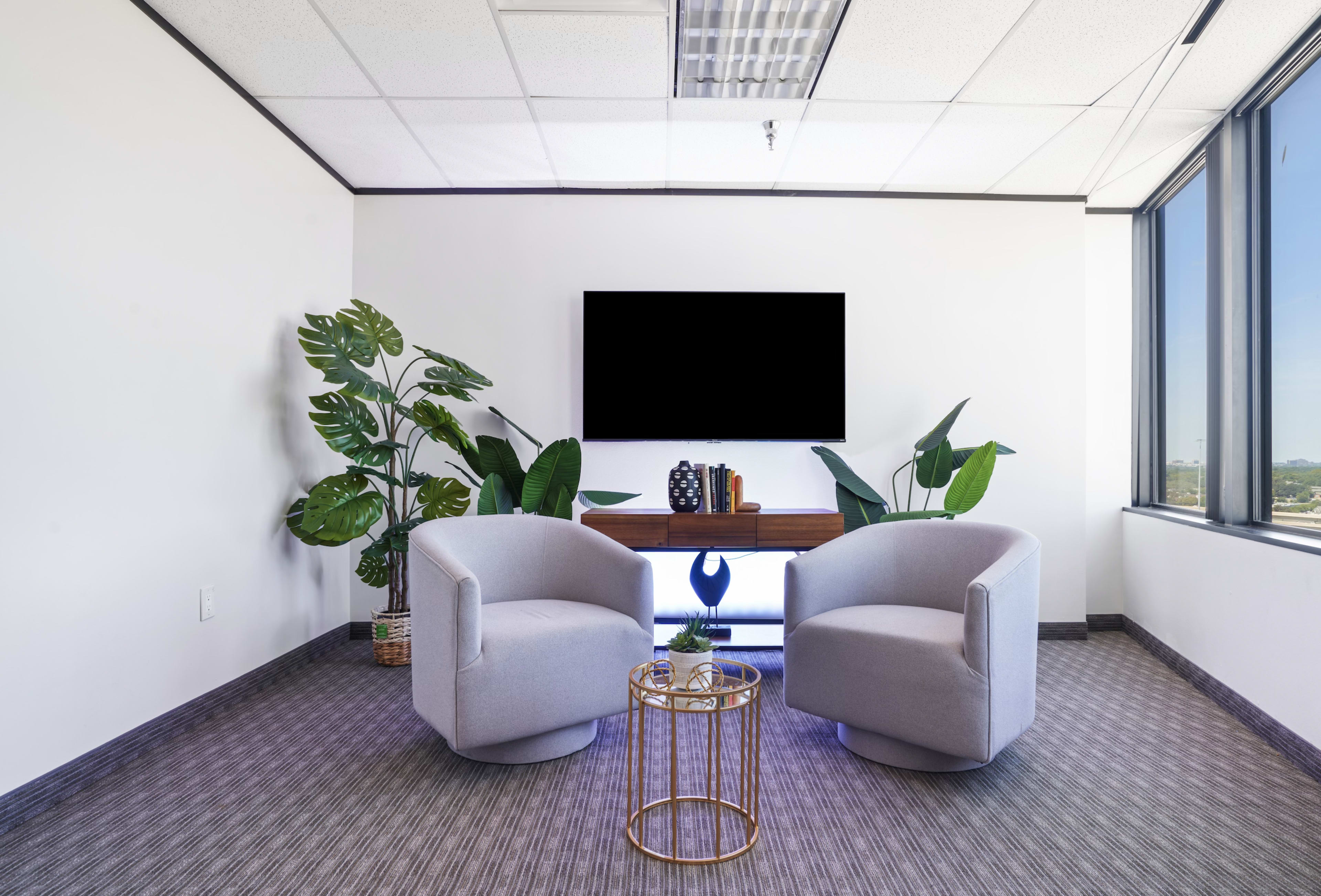 The image shows a modern office space featuring two gray armchairs, a wooden console table with decorative items, and large potted plants beside a wall-mounted television.