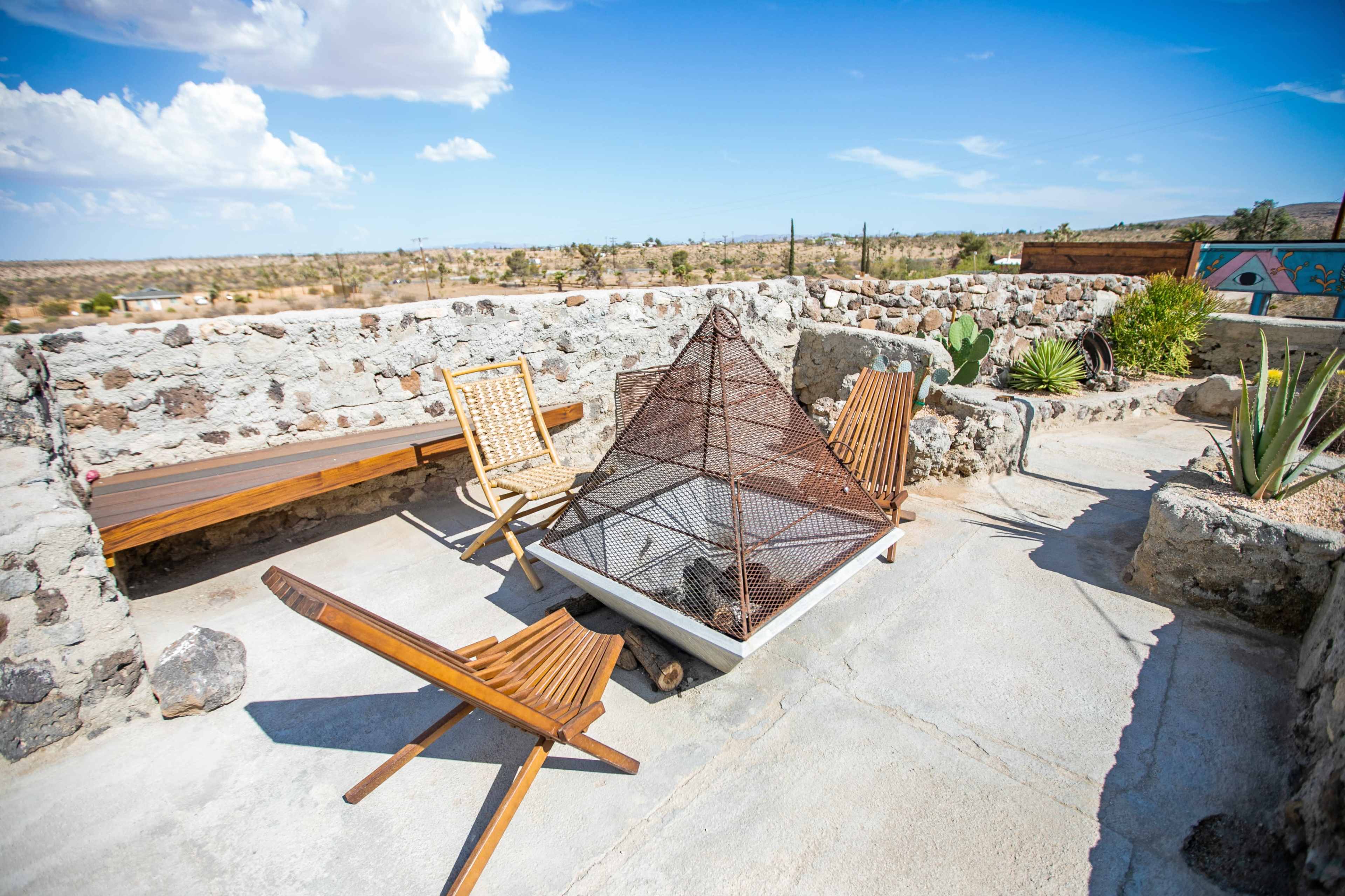 The image shows a rooftop space featuring a metal fire pit, wooden seating, and a stony backdrop against a clear blue sky.