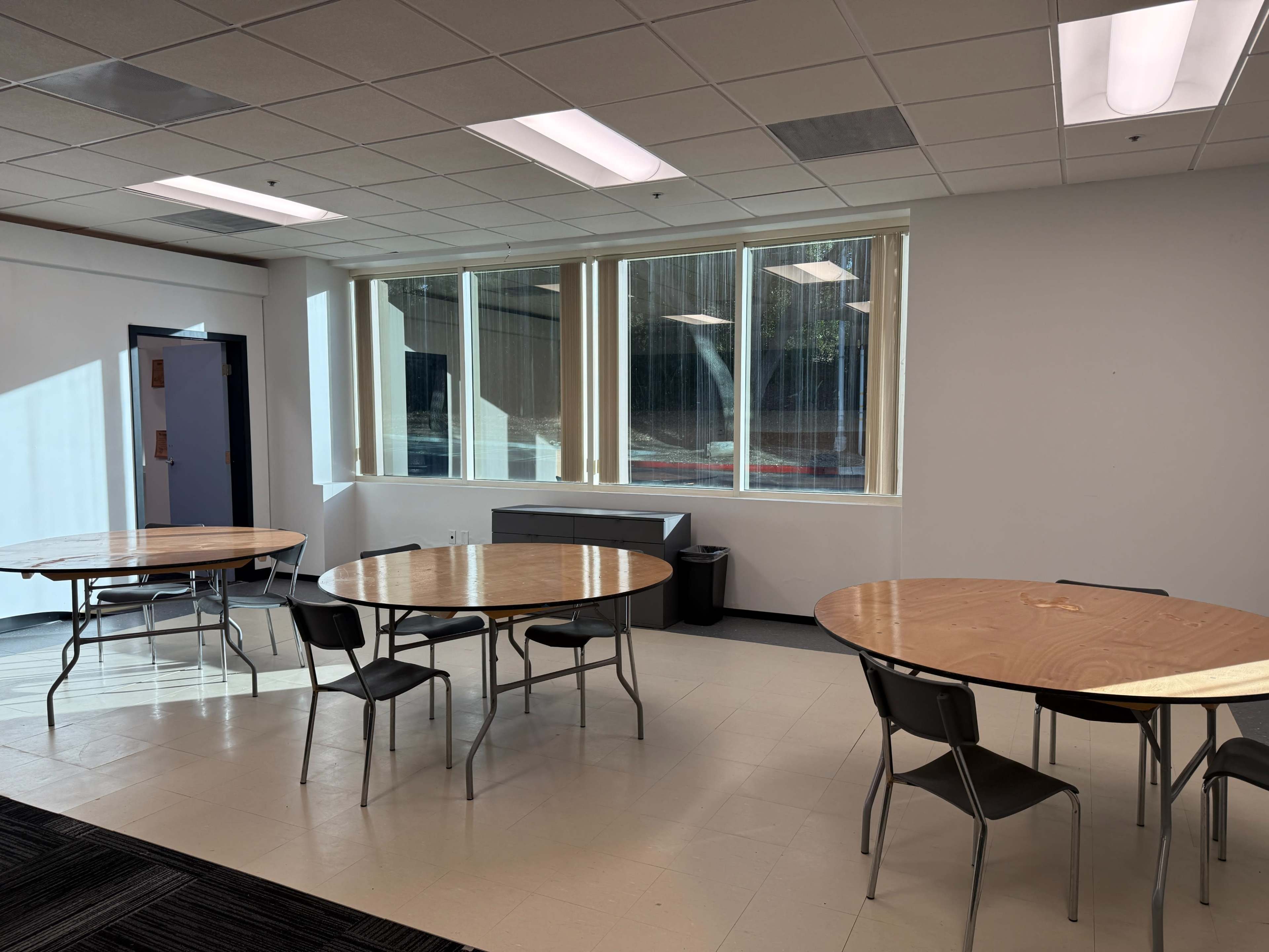 The image shows a room with several round tables and chairs arranged, featuring large windows that let in natural light.