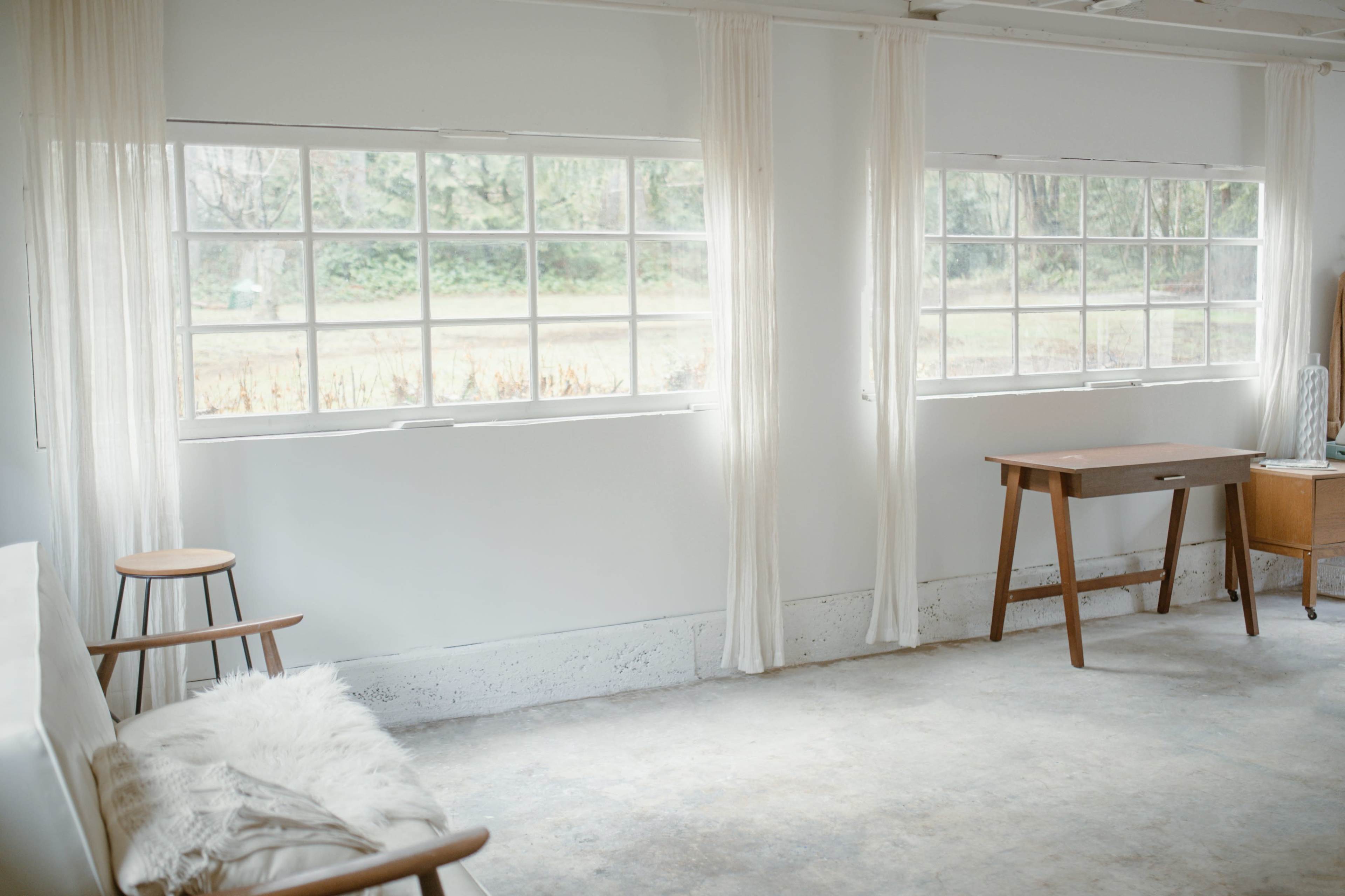 A bright, minimalist room features large windows with sheer curtains, a wooden desk, and a simple chair.