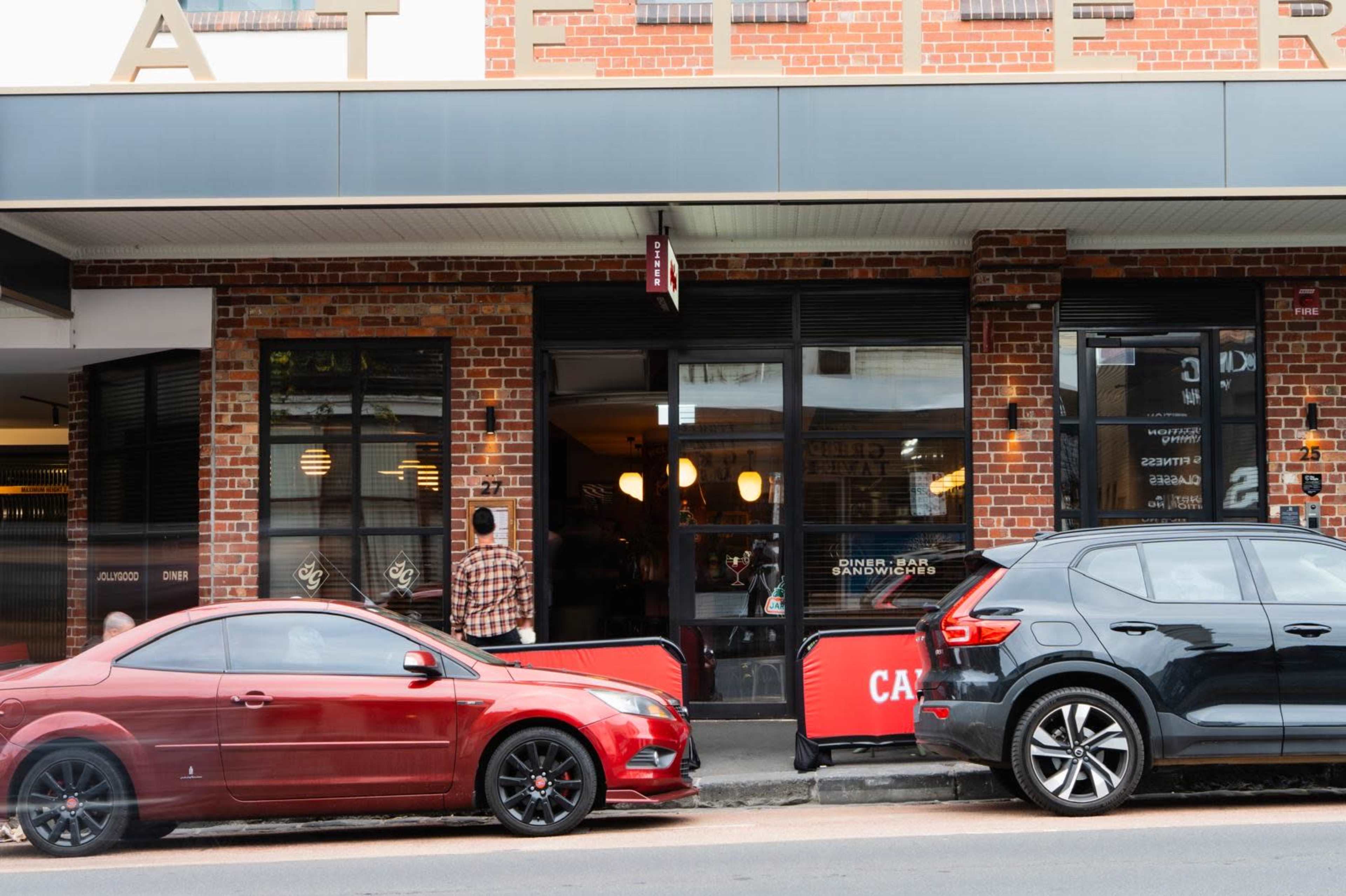 The image shows a street scene with two parked cars—one red and one black—next to a brick building featuring large windows and a cafe sign.