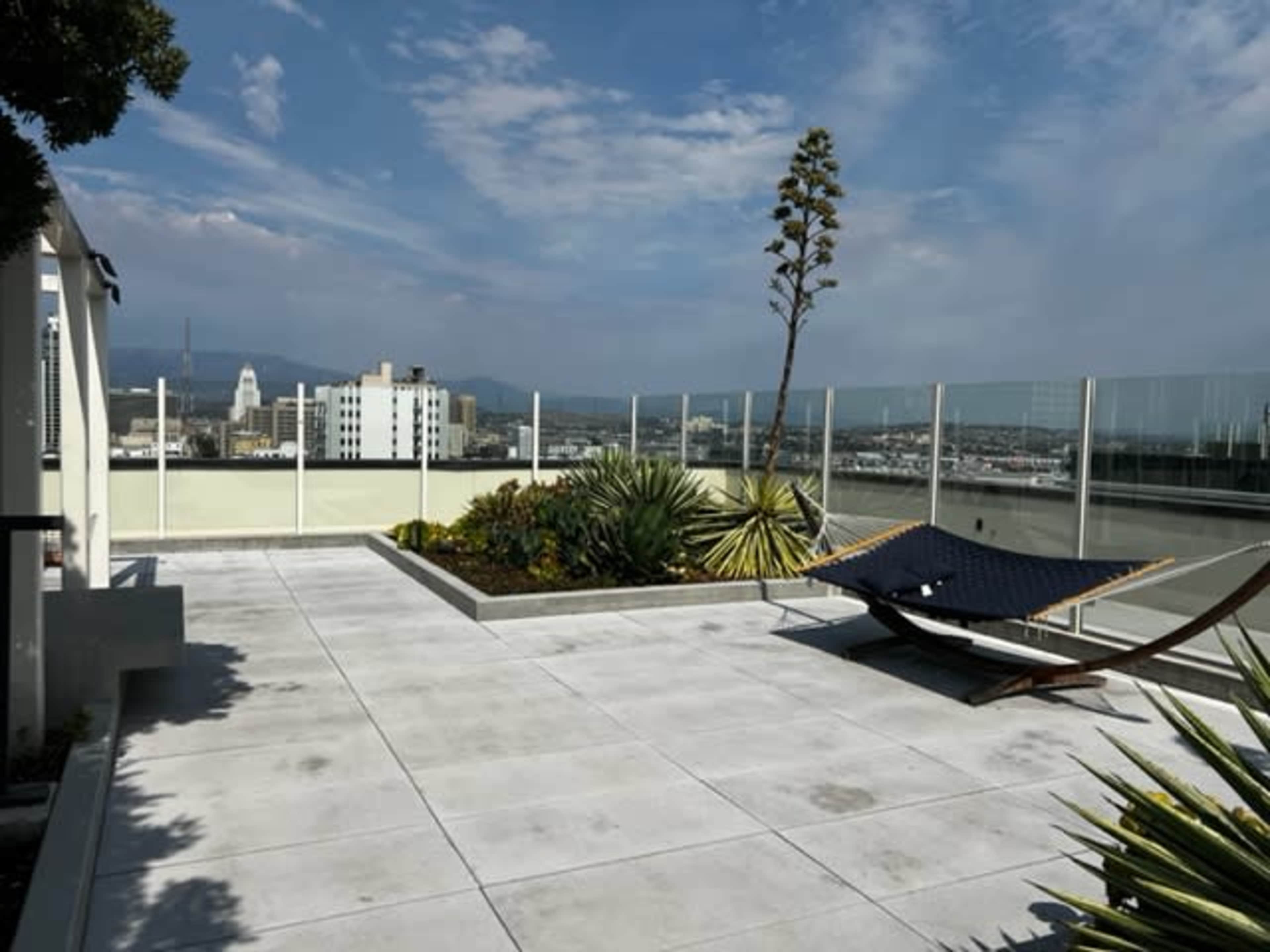 A rooftop terrace features a hammock, potted plants, and a clear view of city buildings under a cloudy sky.