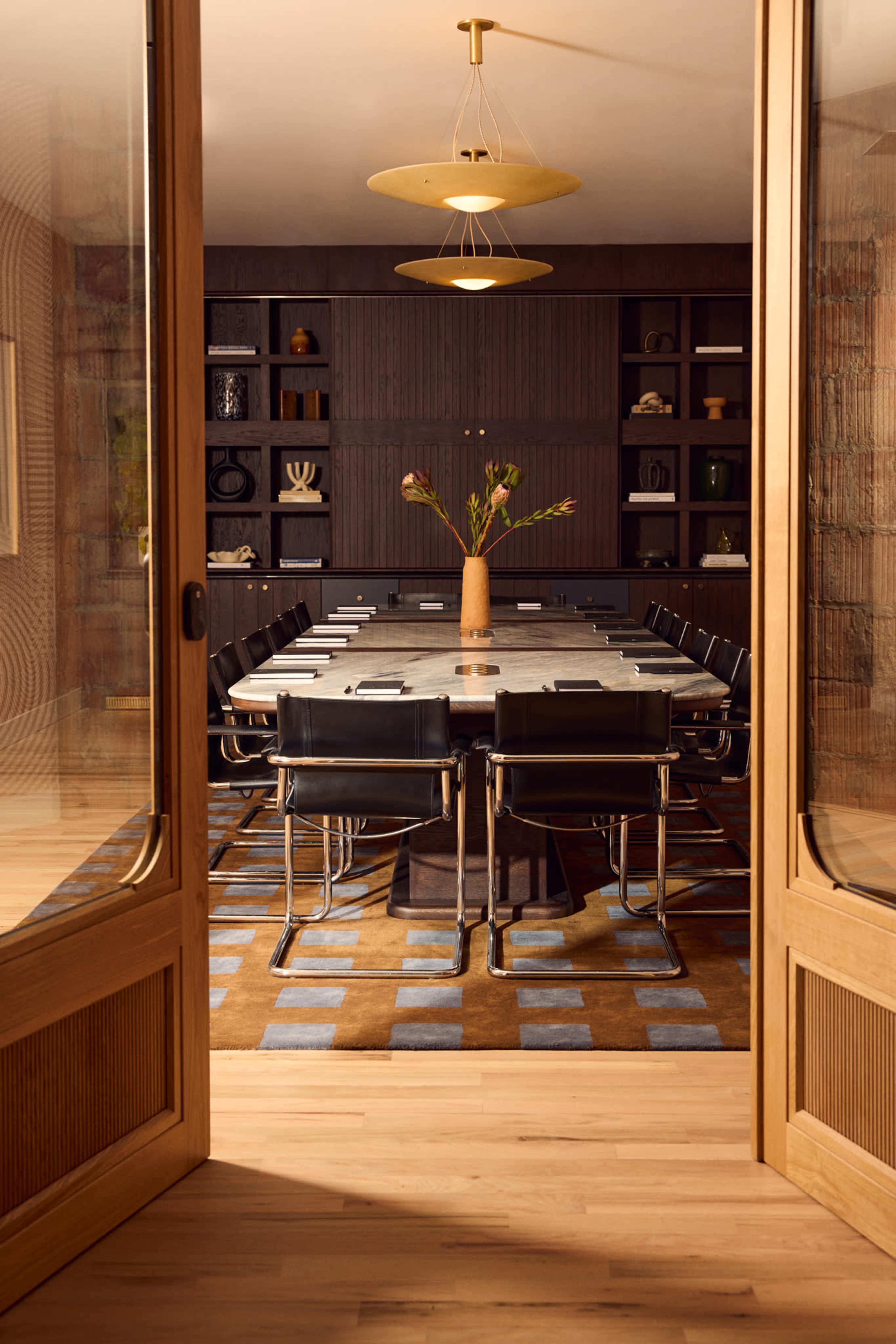A modern conference room features a long table surrounded by black chairs, with a vase of flowers at the center, framed by a wooden doorway.
