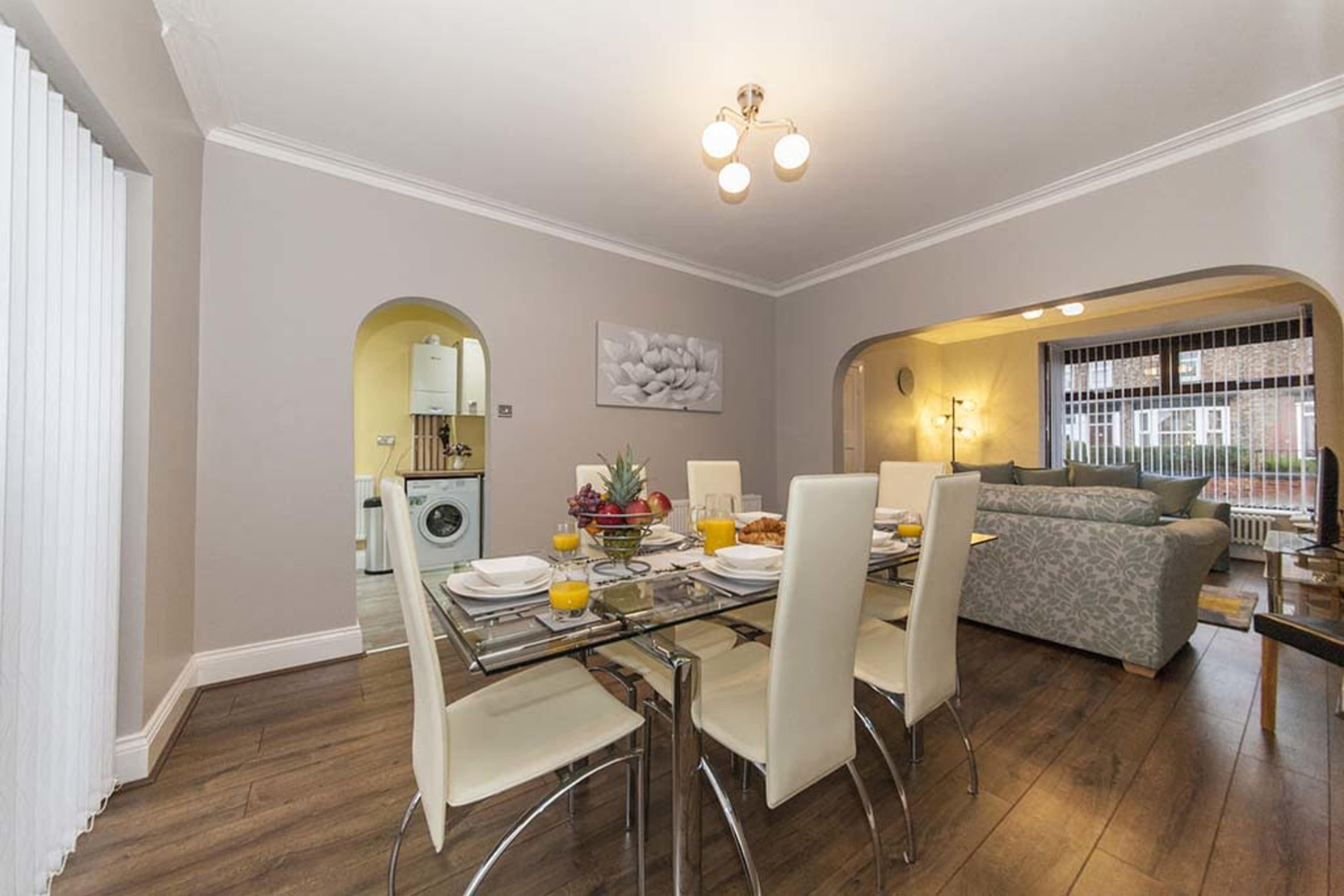 A dining area features a glass table set for a meal, with chairs around it and an open view to a living space beyond.