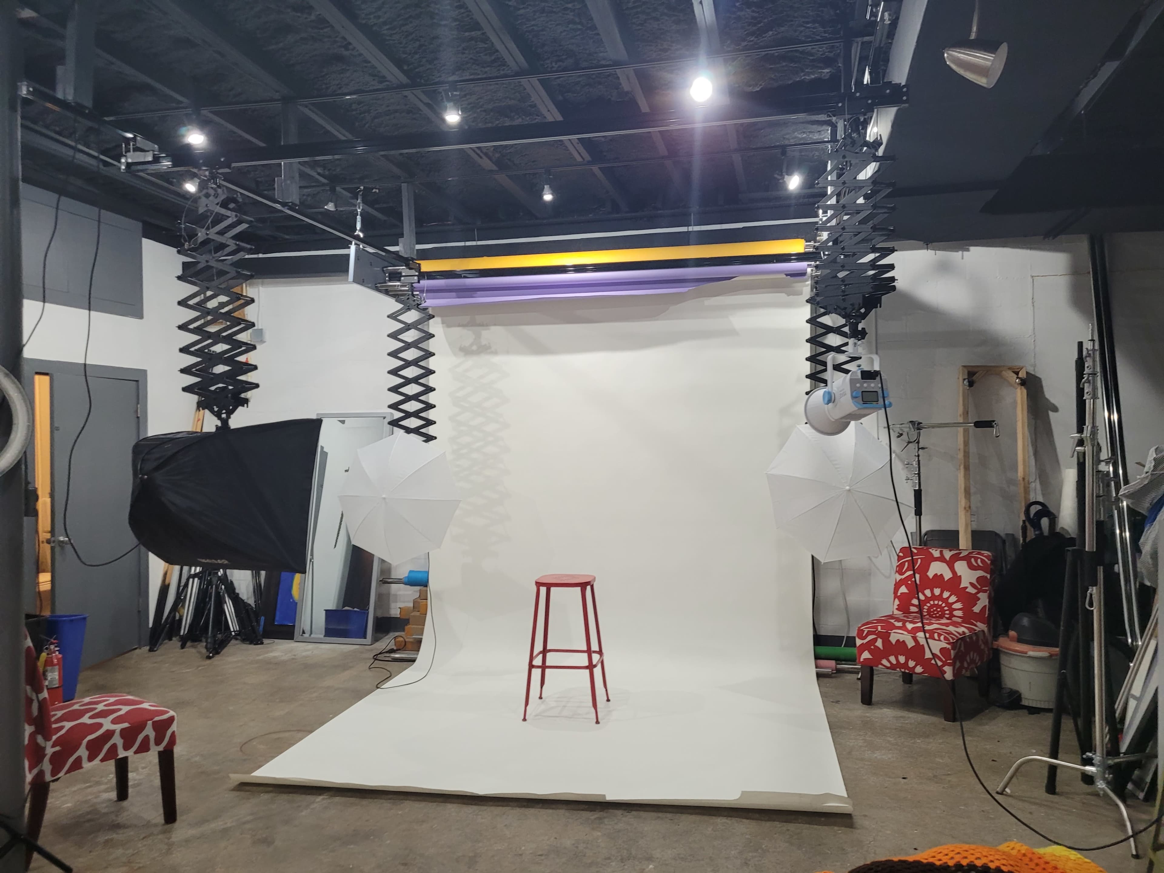 The image shows a photography studio equipped with a blank backdrop, a red stool, and lighting umbrellas set up for photo shoots.