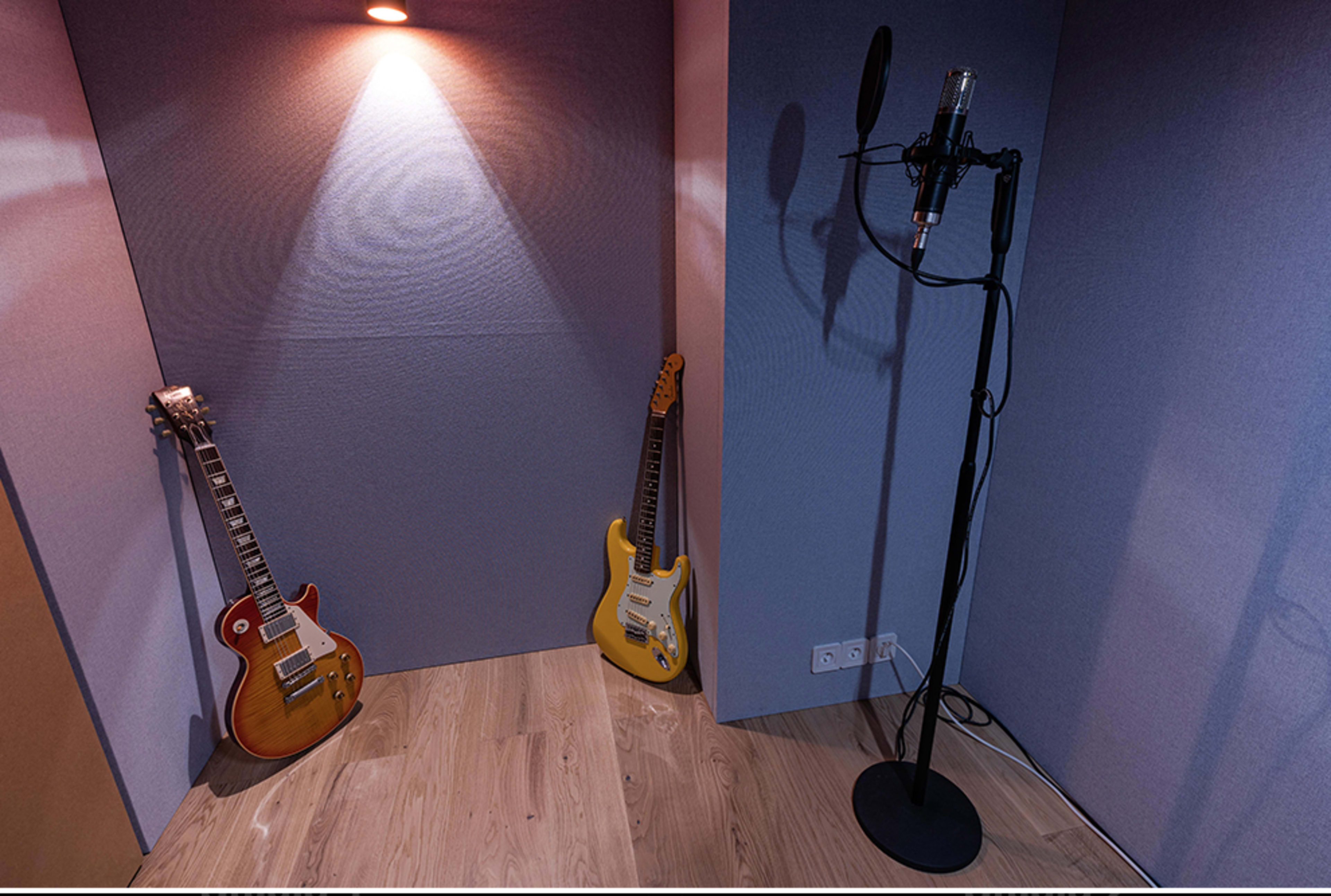 A small recording booth featuring two electric guitars positioned against the wall and a microphone on a stand.