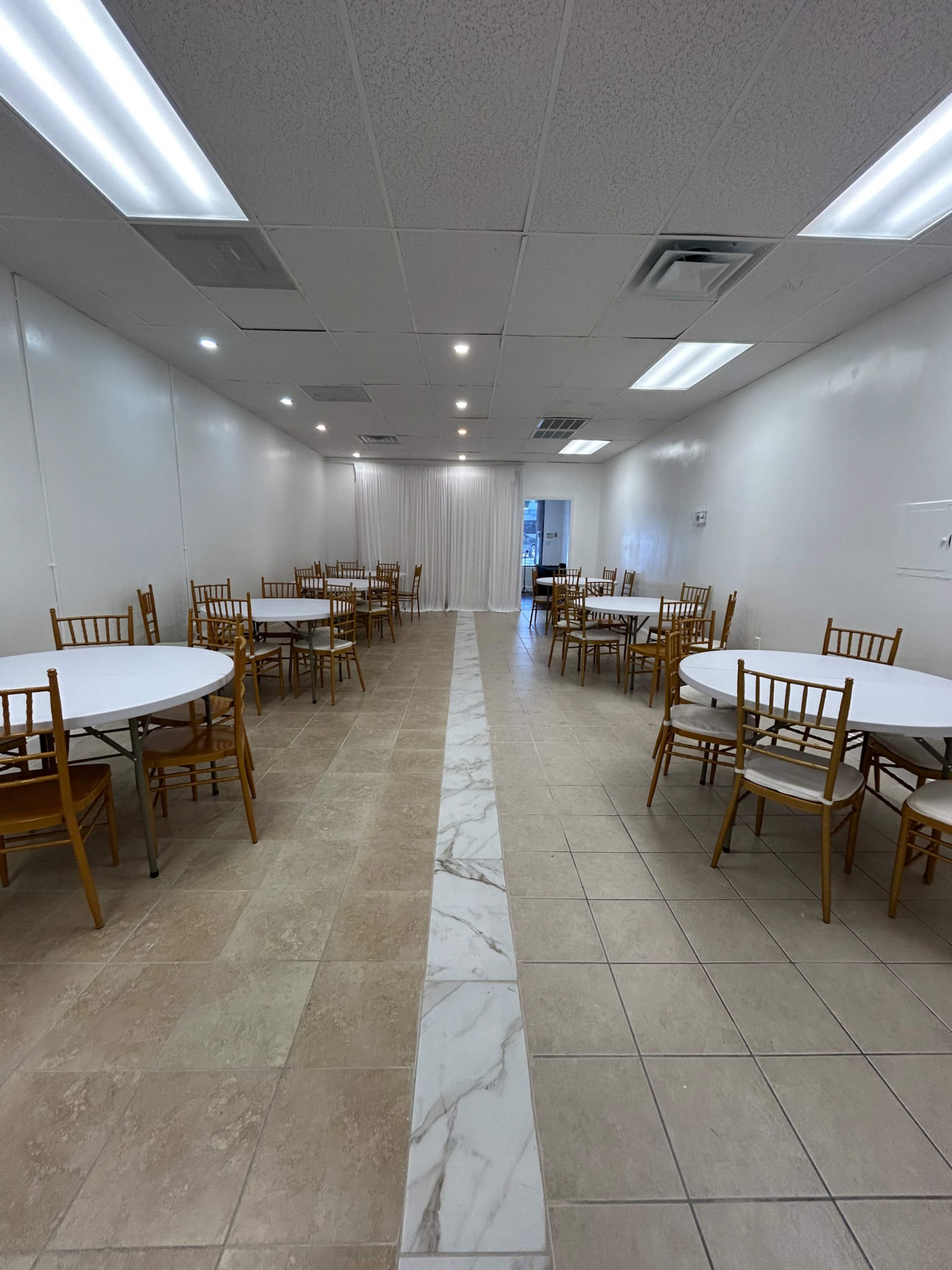 A spacious room features several round tables with wooden chairs arranged along a central strip of marble flooring.