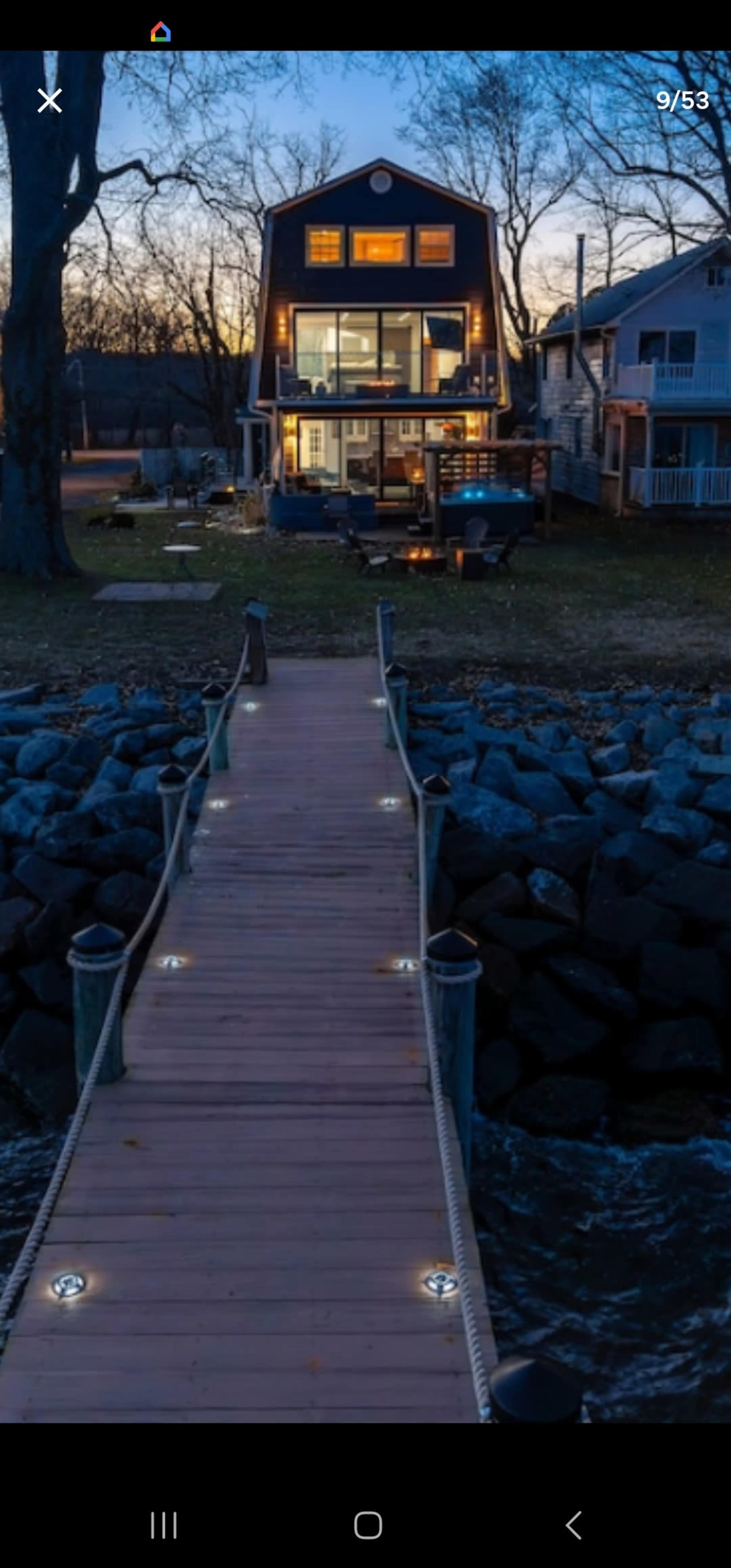 A wooden dock lined with lights leads to a modern house with large windows, situated by the water's edge at dusk.