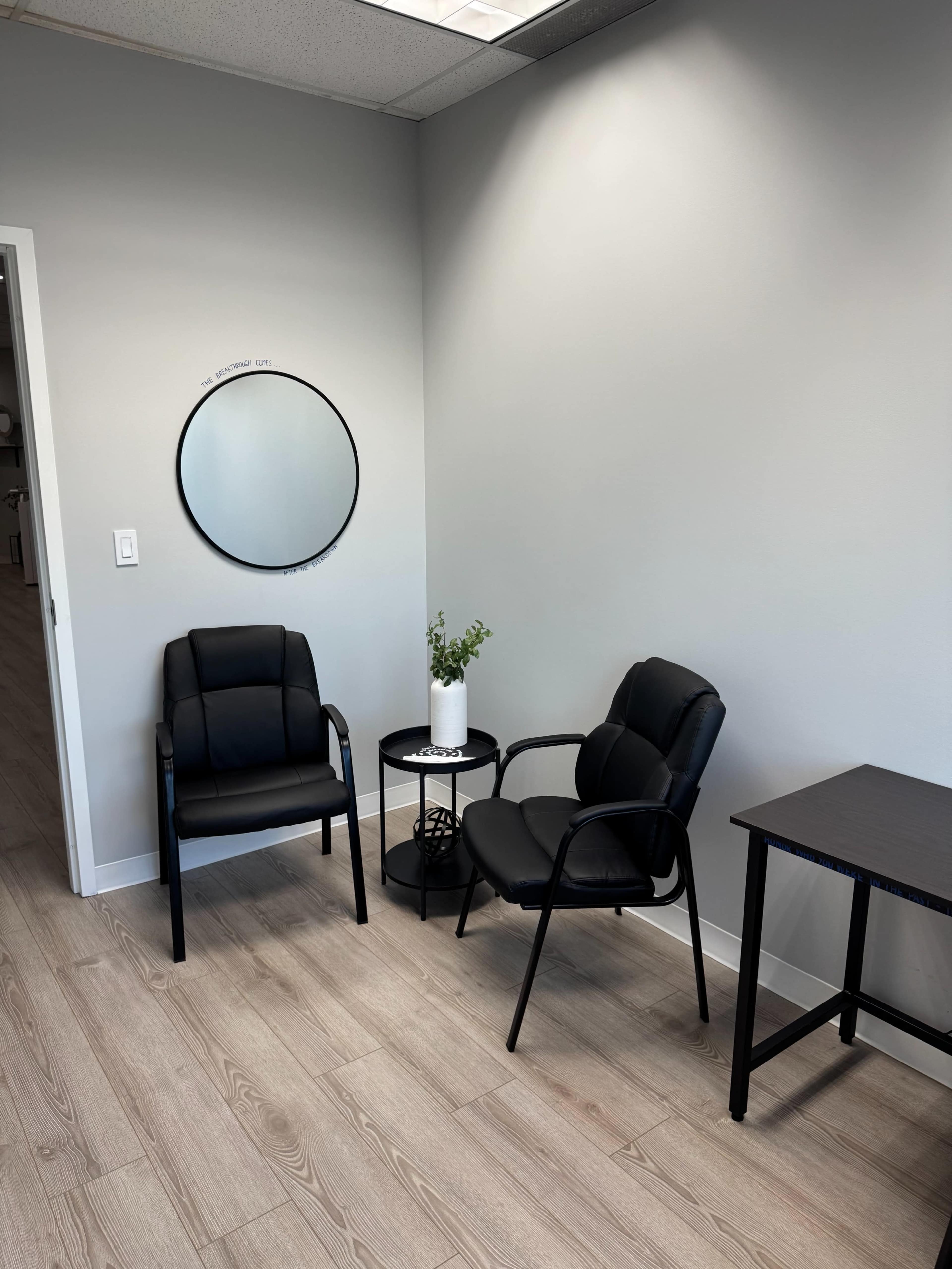 The image shows a small waiting area with two black chairs, a round mirror on the wall, a small table, and a white planter with a plant.