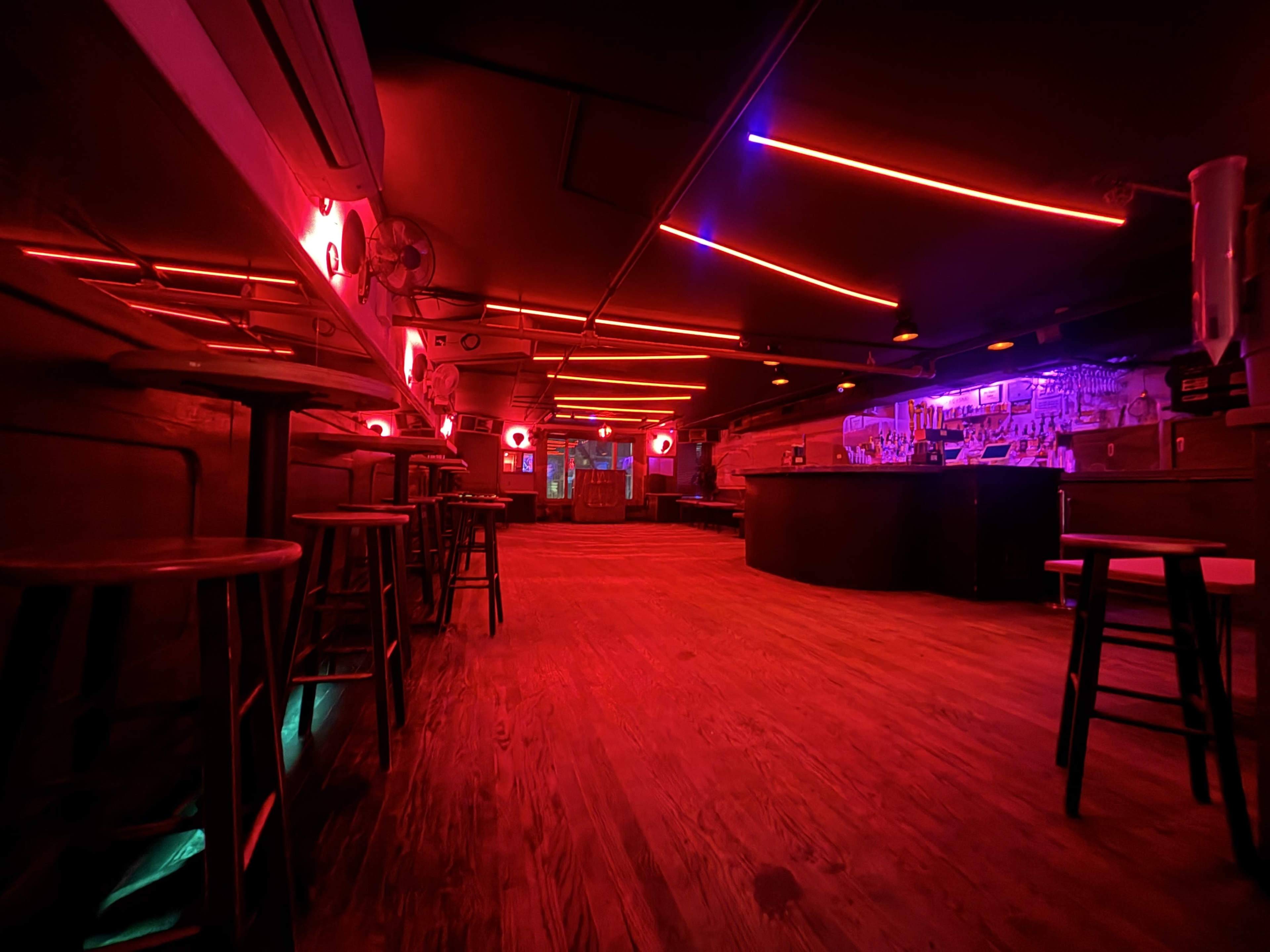 The image shows an empty bar interior illuminated by red neon lights, with wooden floors and stools arranged along the counter.