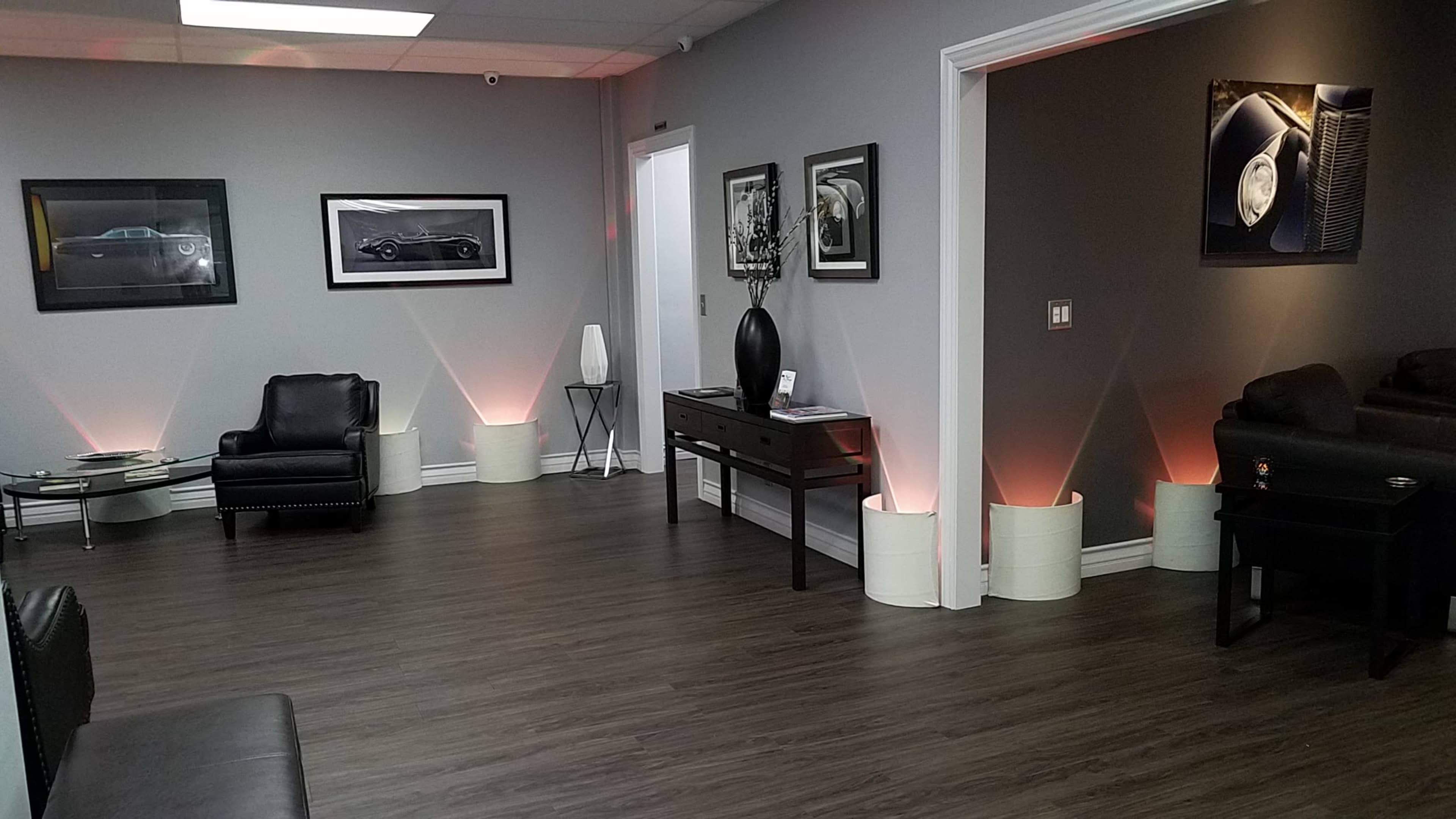 The image shows a sleek, modern interior space with dark wood flooring, framed black-and-white car photographs on the walls, and minimalist furniture.