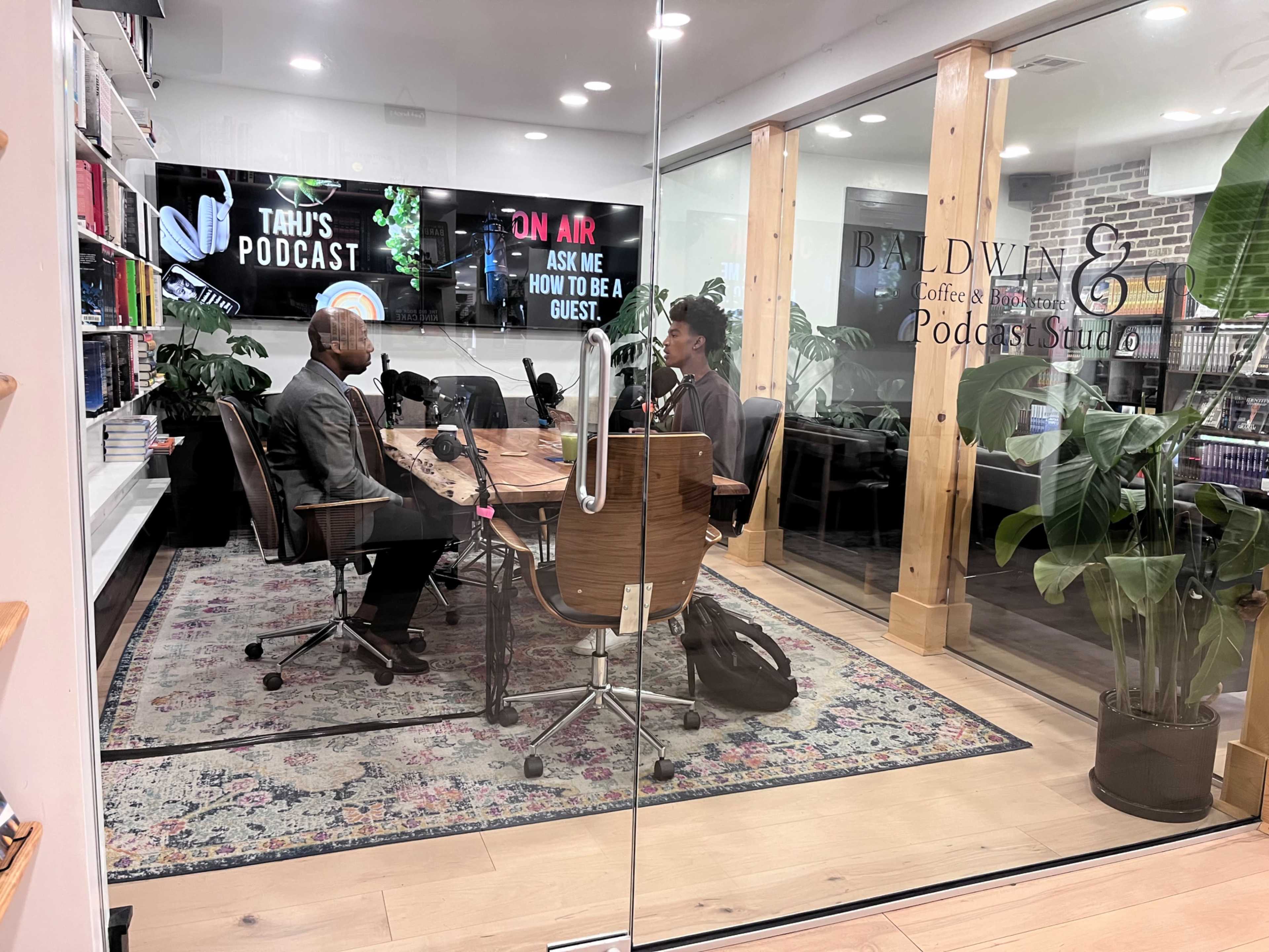 A man and a woman sit at a table with microphones in a podcast studio, surrounded by bookshelves and plants, while a screen displays "ON AIR."