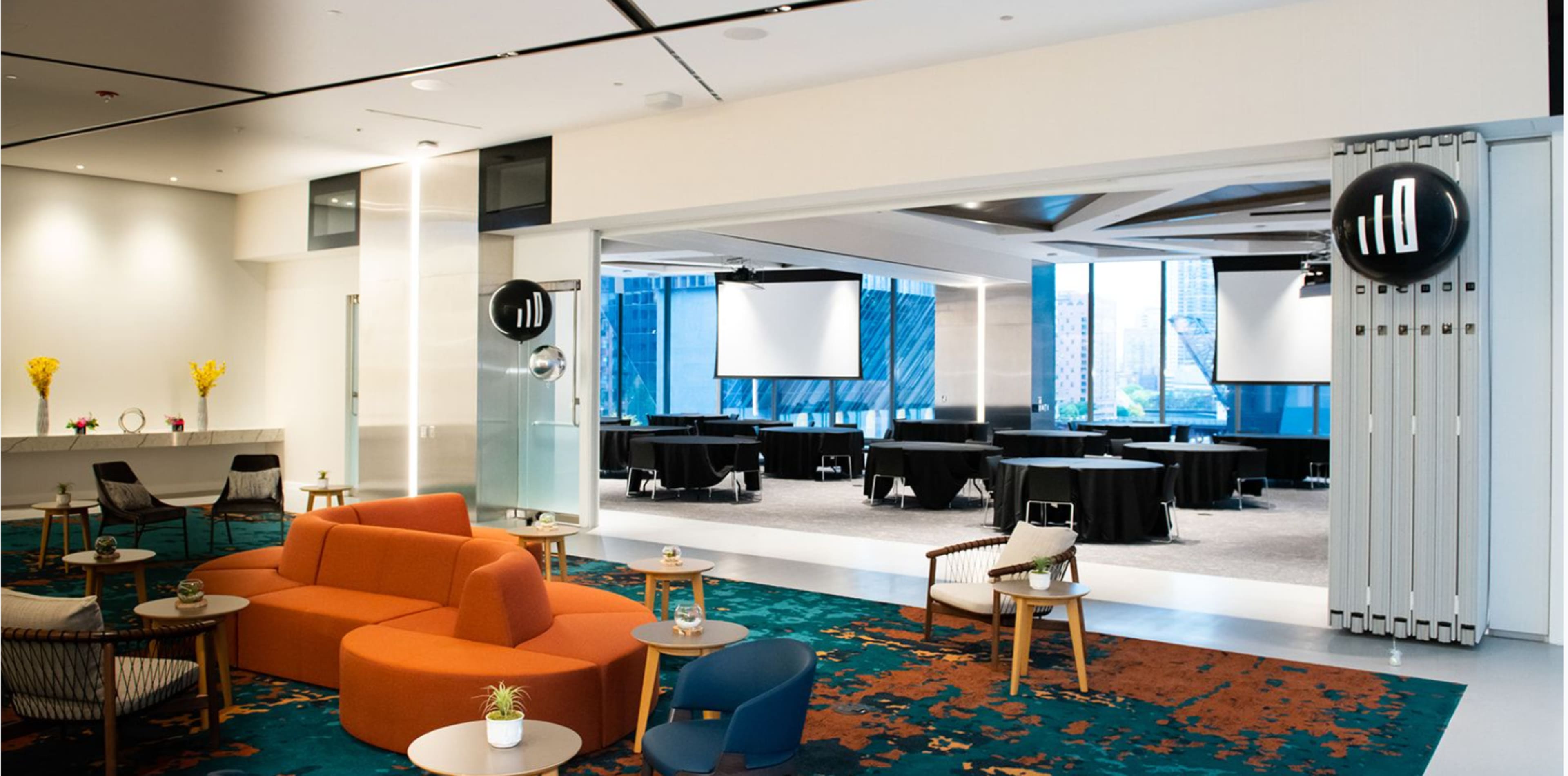 The image shows a modern event space with a lounge area featuring an orange sofa and several seating arrangements, alongside a conference area with black tables and projectors visible through large windows.