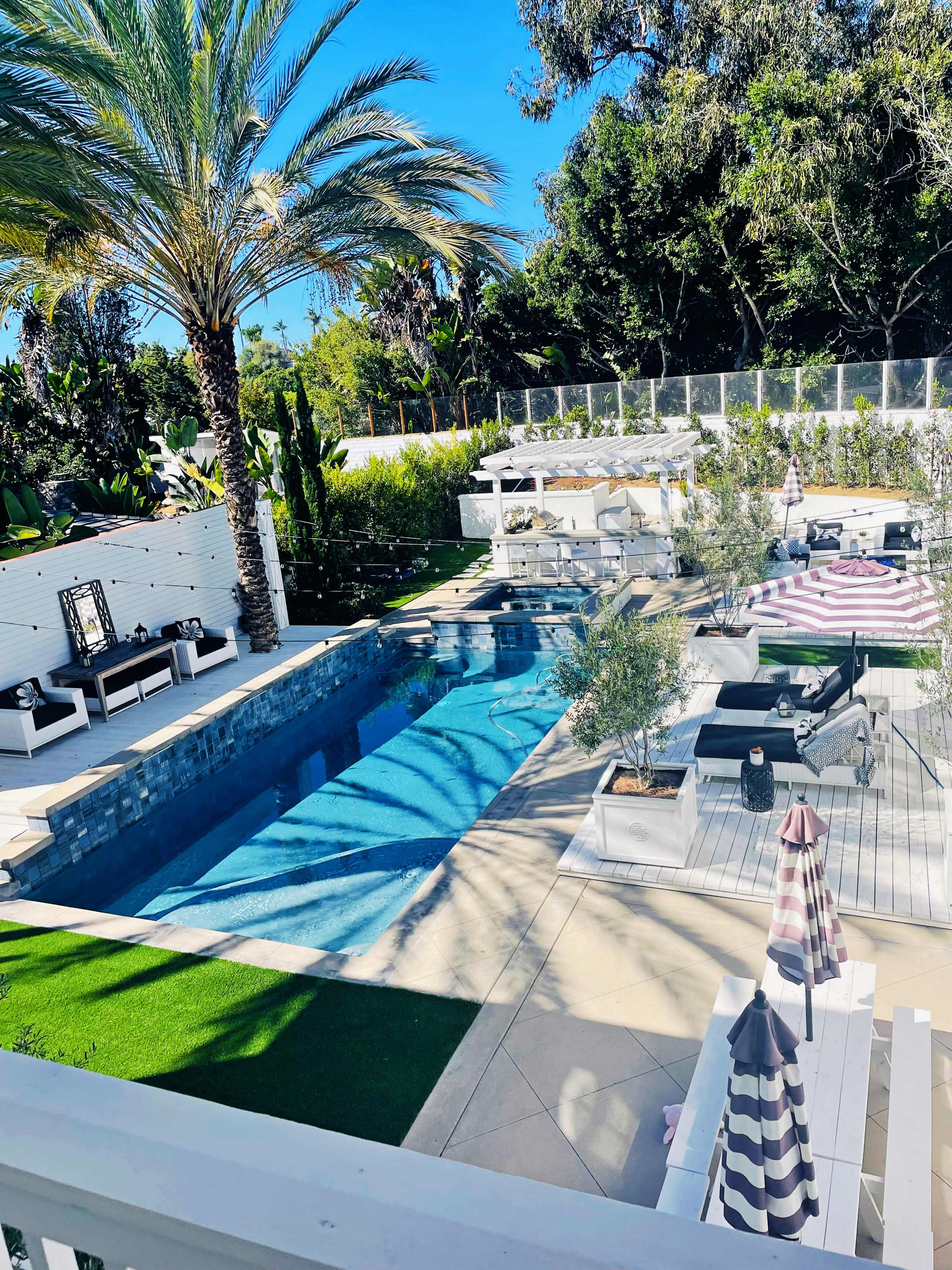 The image shows a spacious outdoor area featuring a rectangular swimming pool surrounded by lounge furniture, sunbeds, and tropical landscaping.