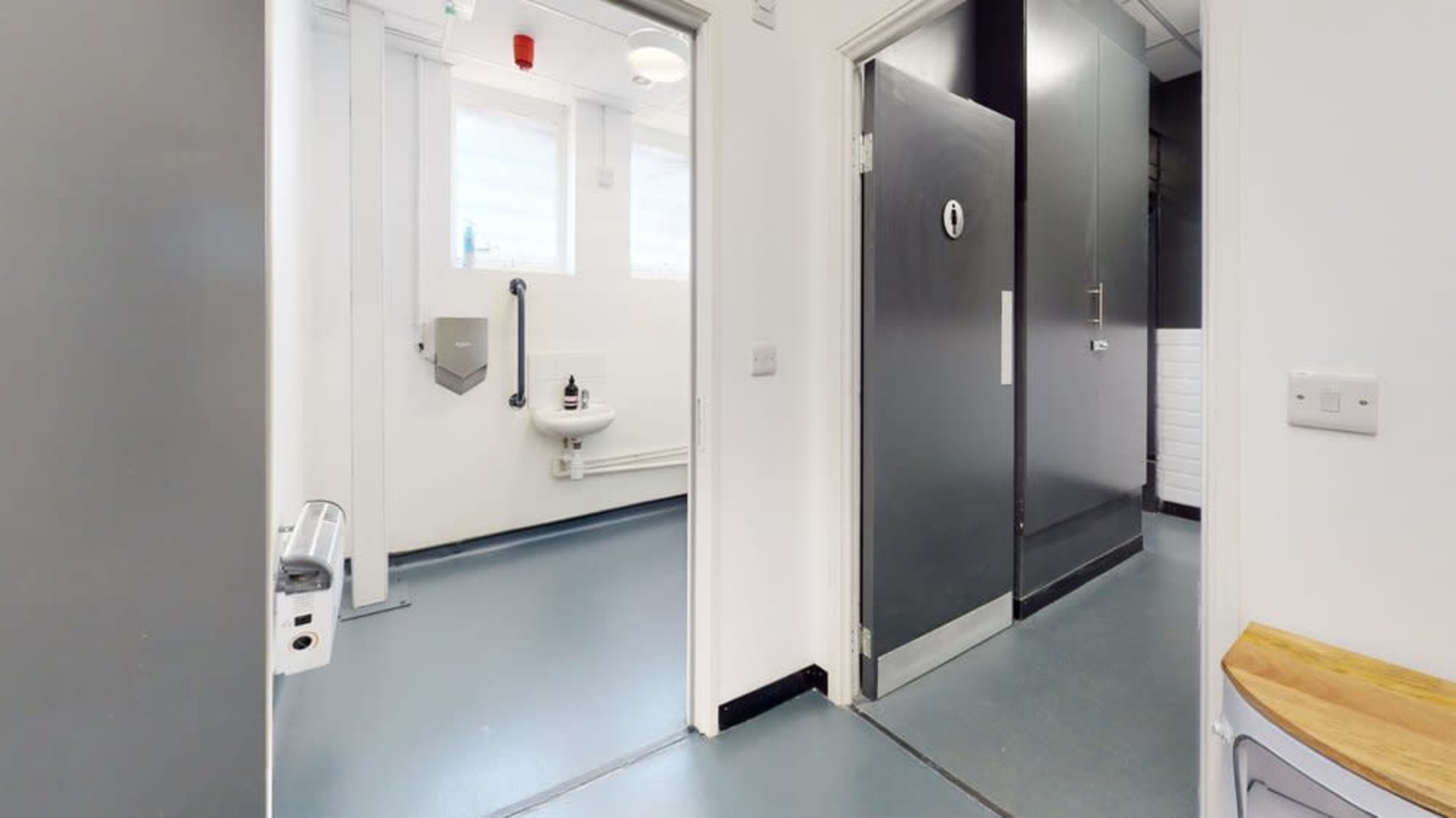 An interior hallway leading to two doors, with one door open to a small restroom area featuring a sink and a black wall.