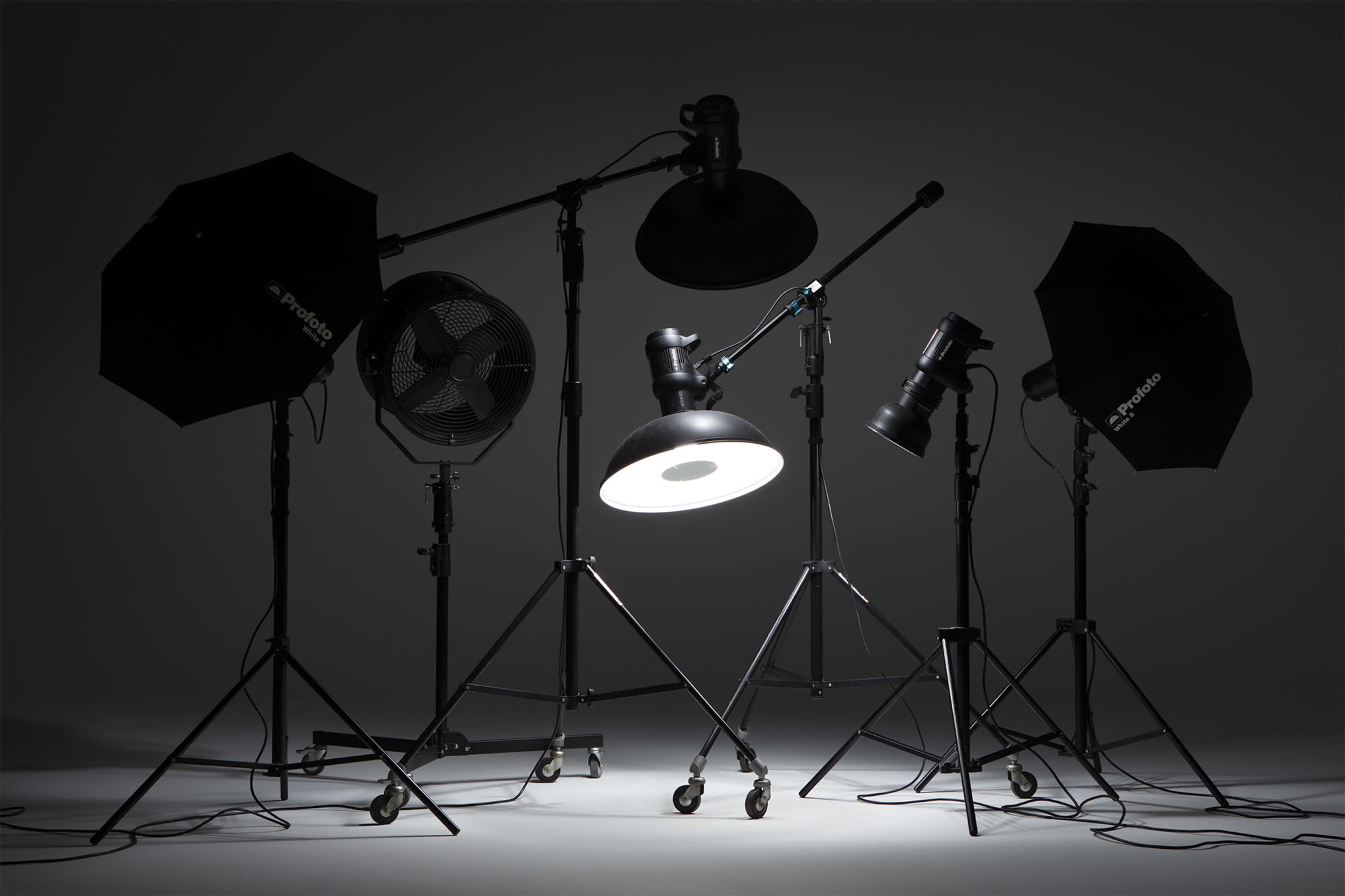 Various photography studio lighting equipment, including soft boxes, a fan, and spotlights on adjustable stands against a dark background.