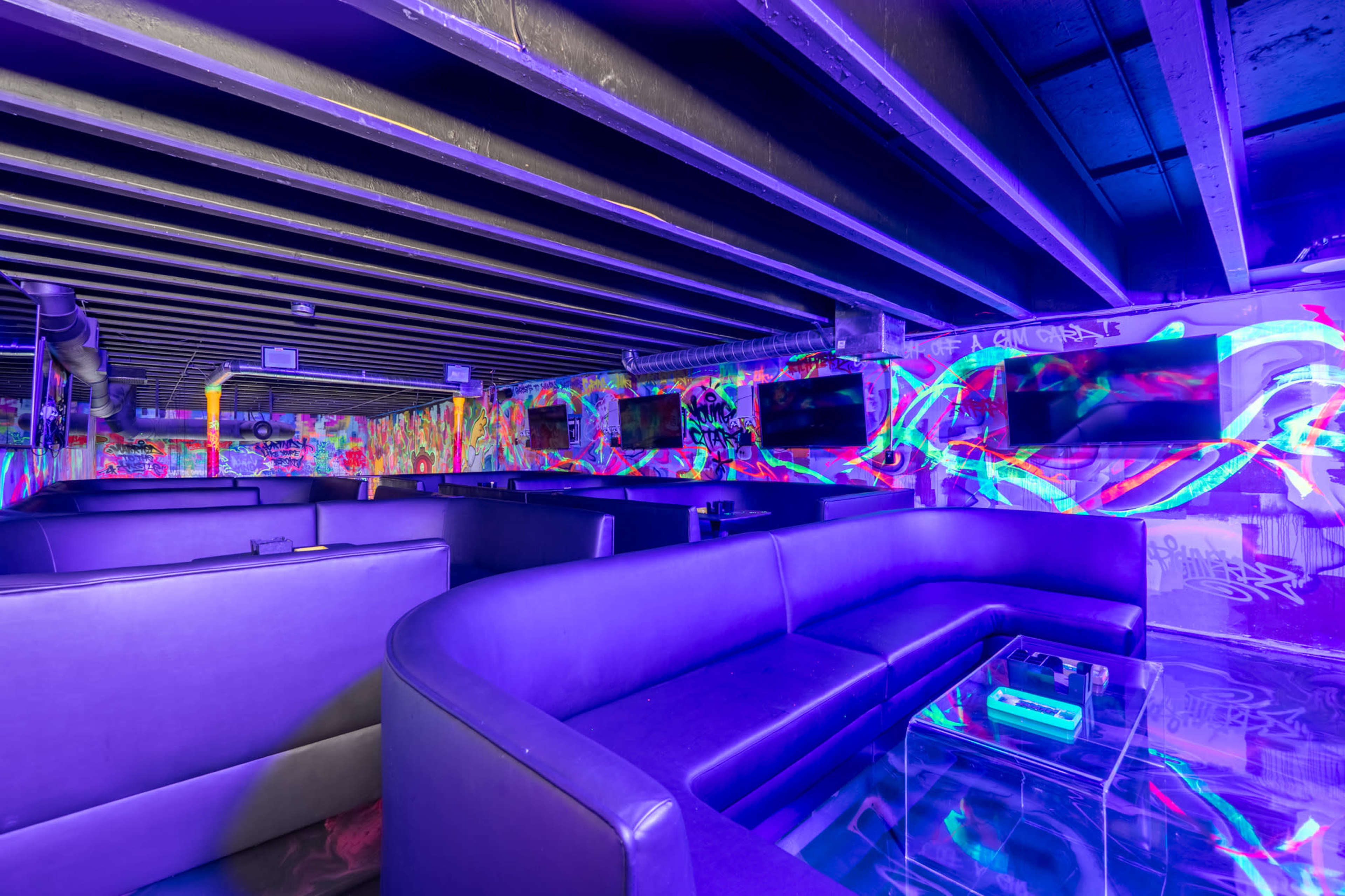 The image shows a dimly lit lounge with colorful graffiti on the walls and large, black leather sofas arranged around a glass coffee table.