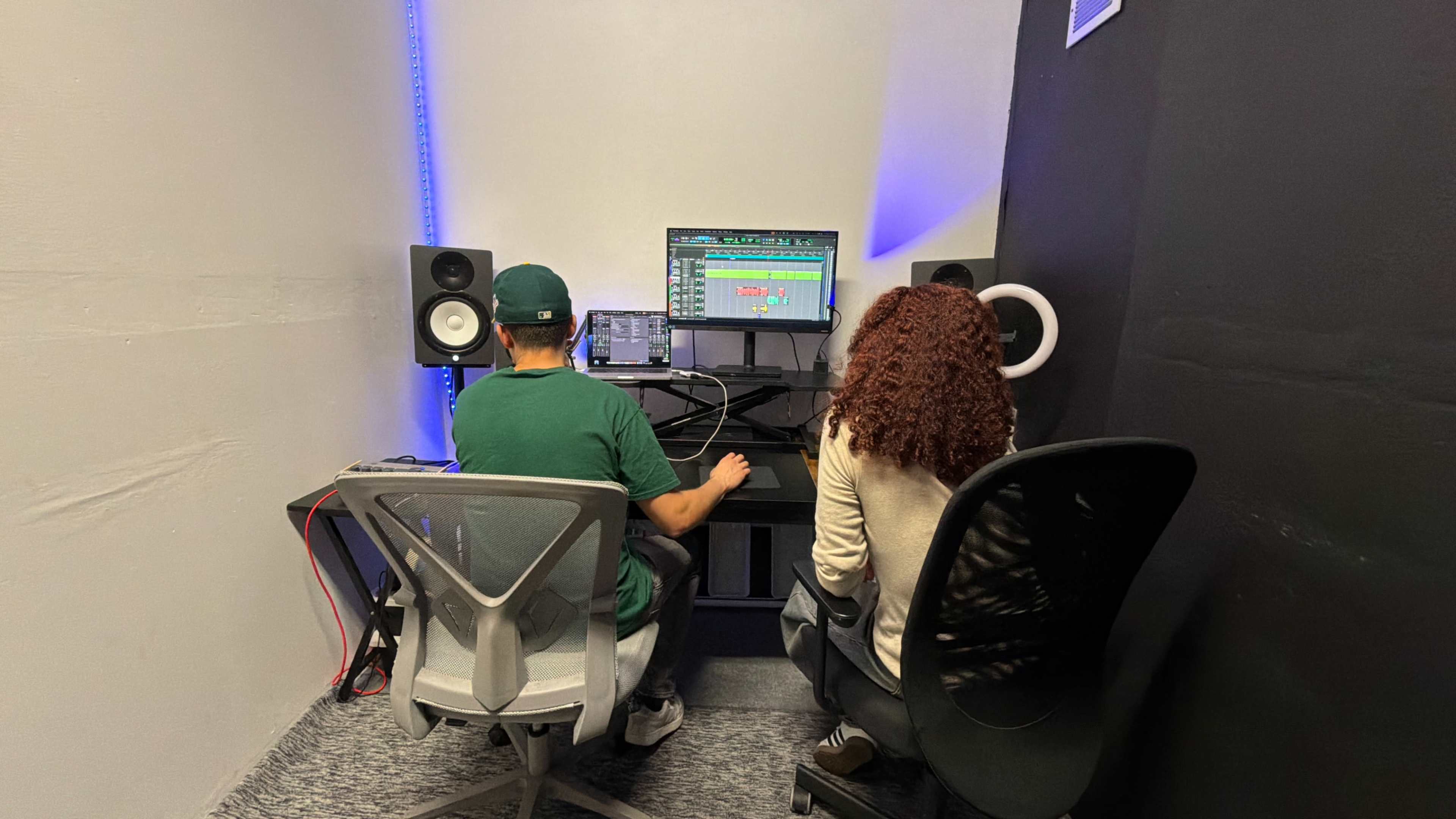 Two individuals are seated at a desk in a recording studio, working on music software with studio monitors on either side.