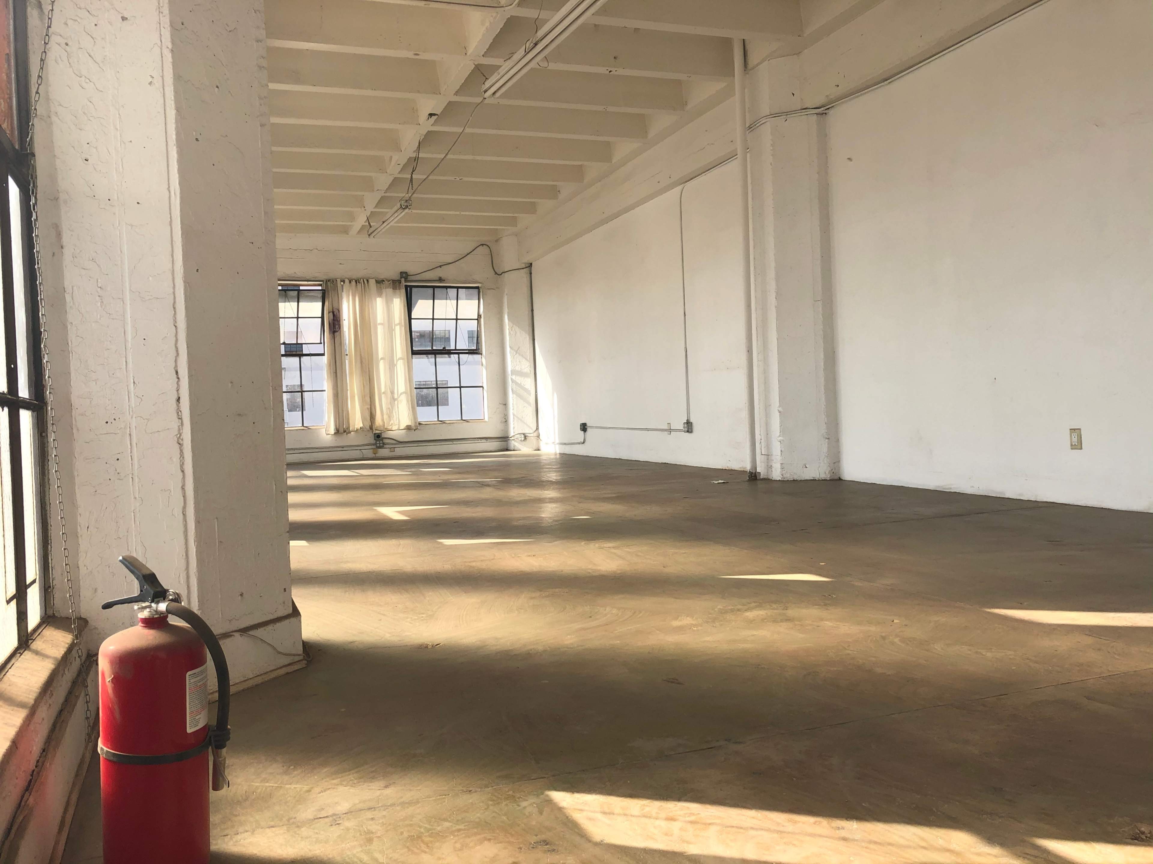 An empty room with polished concrete floors, large windows with sheer curtains, and a fire extinguisher on the floor.