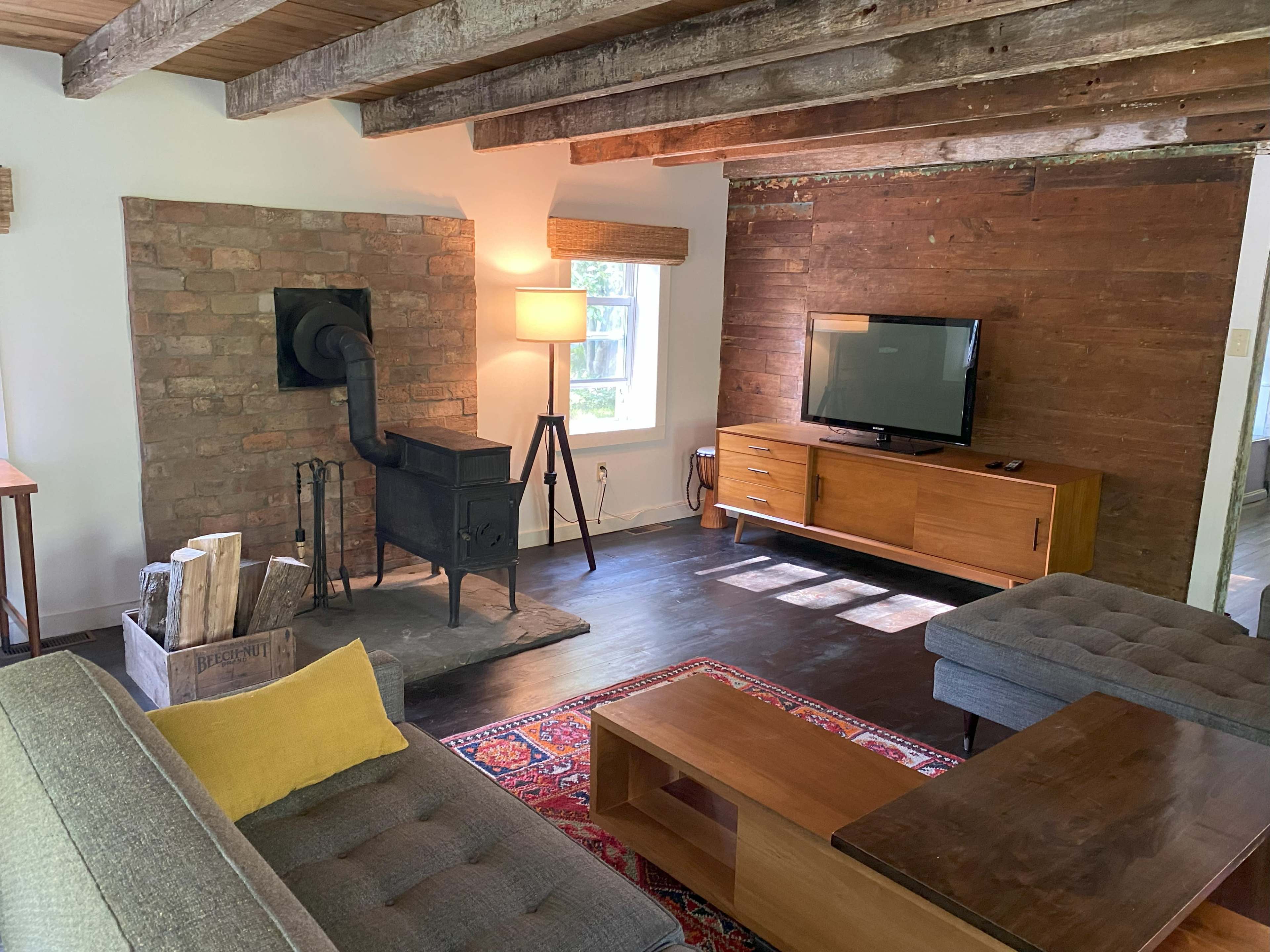 The image shows a cozy living room featuring a wood stove, a television on a wooden cabinet, and two gray sofas arranged around a coffee table.