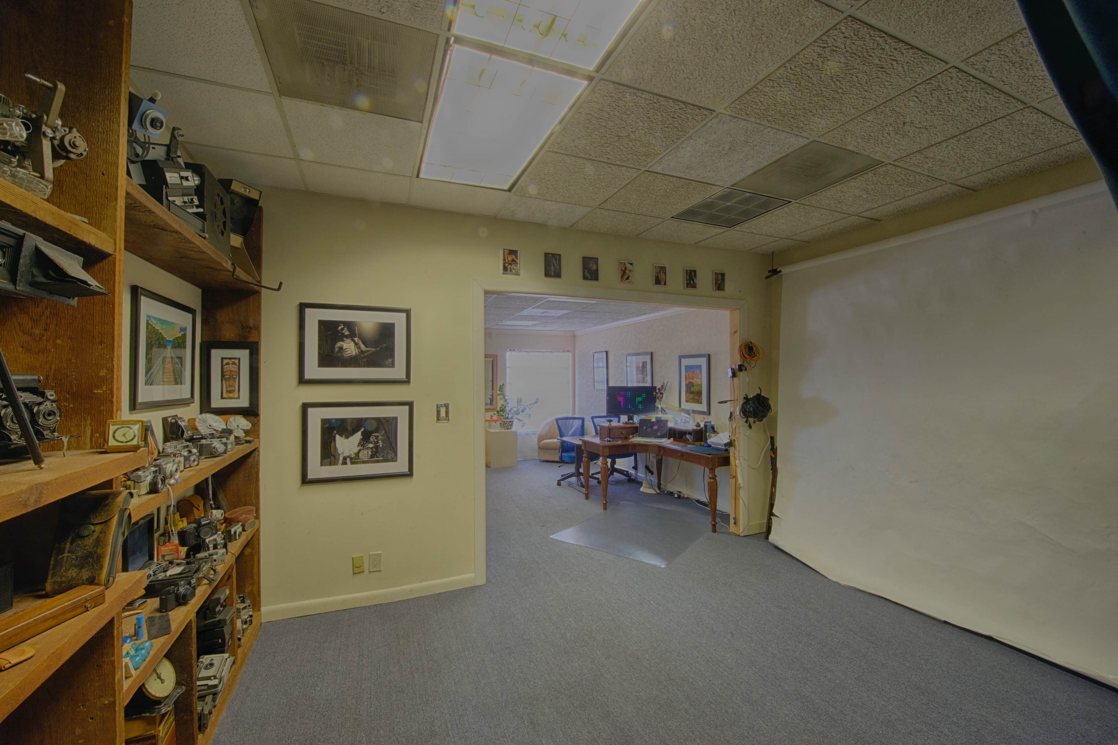The image shows a room with wooden shelves displaying cameras, framed photographs on the walls, and an adjoining area with a desk and chairs visible in the background.