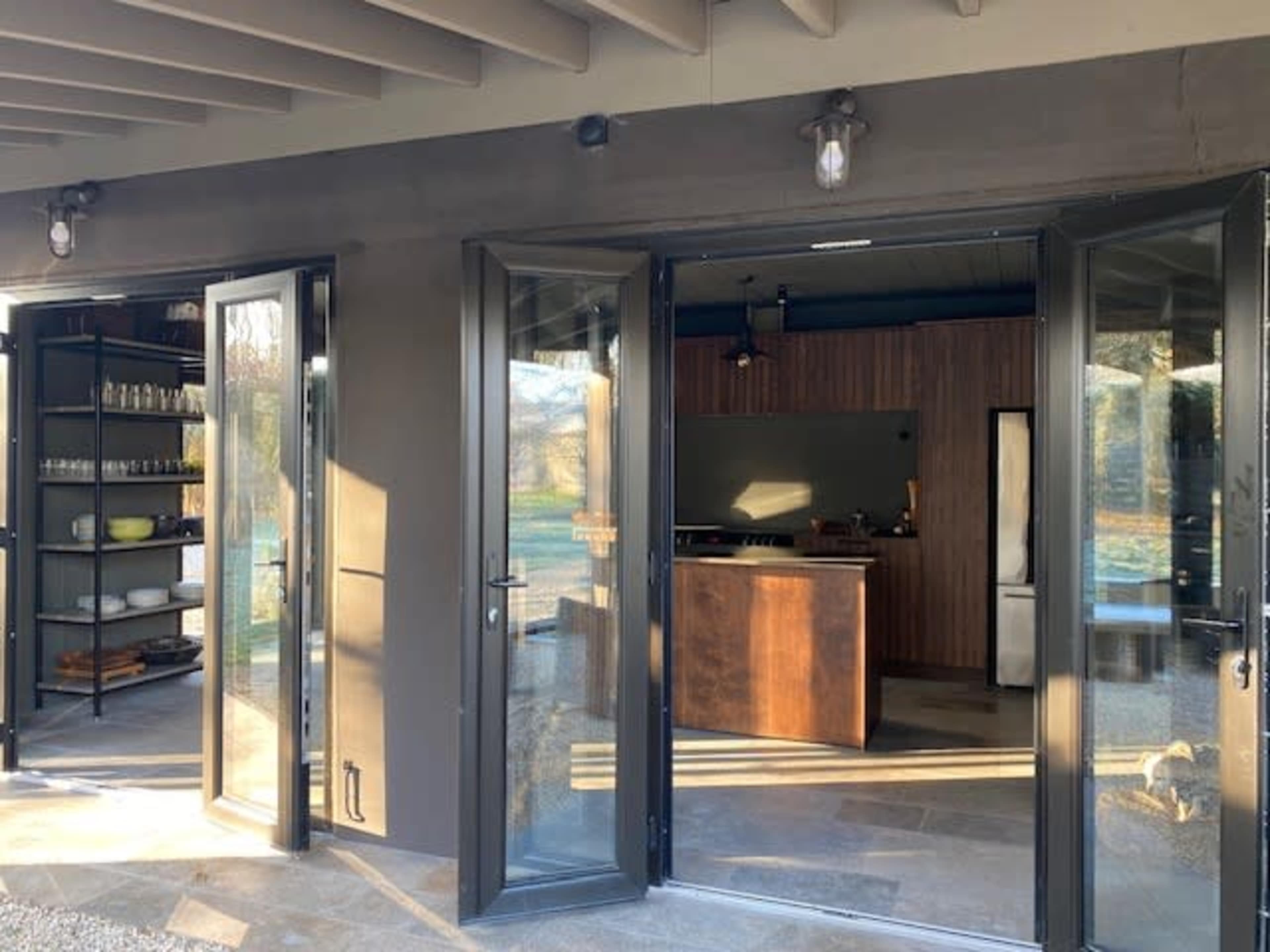 The image shows a modern kitchen with open doors leading to an outdoor area, featuring shelving with various items on the left and a wooden kitchen island inside.