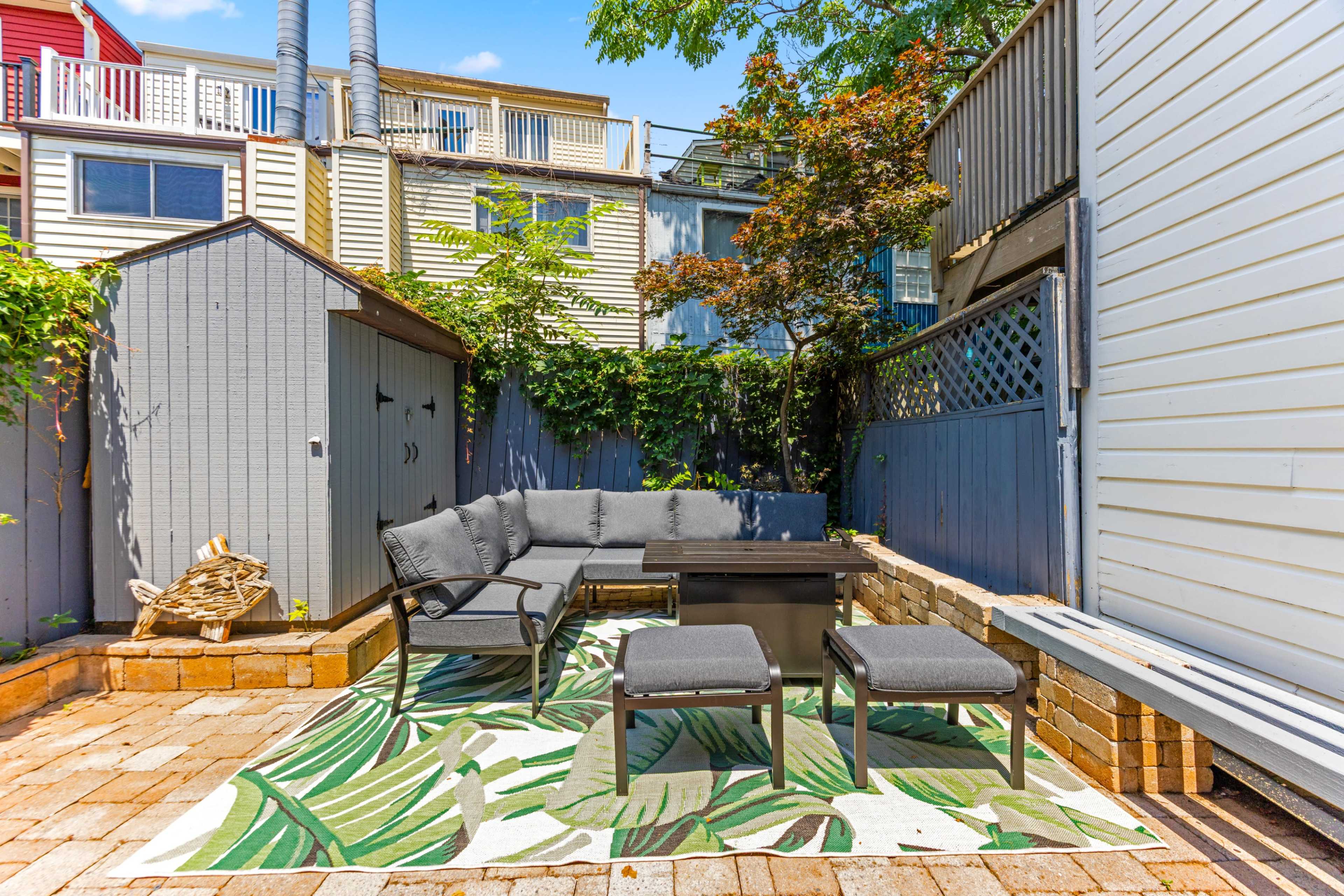 A small outdoor patio features a gray seating arrangement, a coffee table, and a decorative rug, surrounded by wooden fences and sheds.