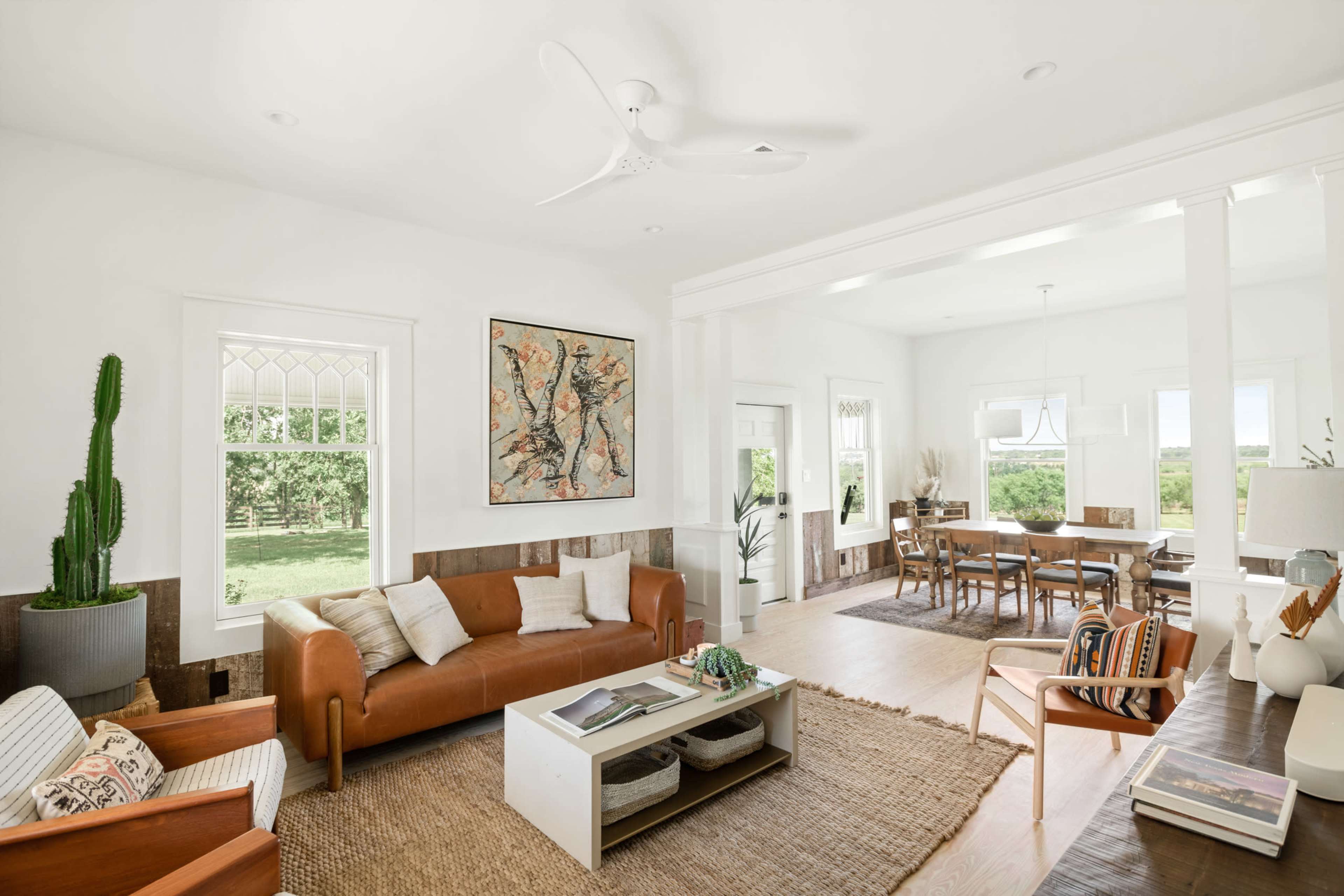 A bright, modern living space features a brown leather sofa, a coffee table, and a large cactus, with natural light streaming in from windows overlooking a green landscape.